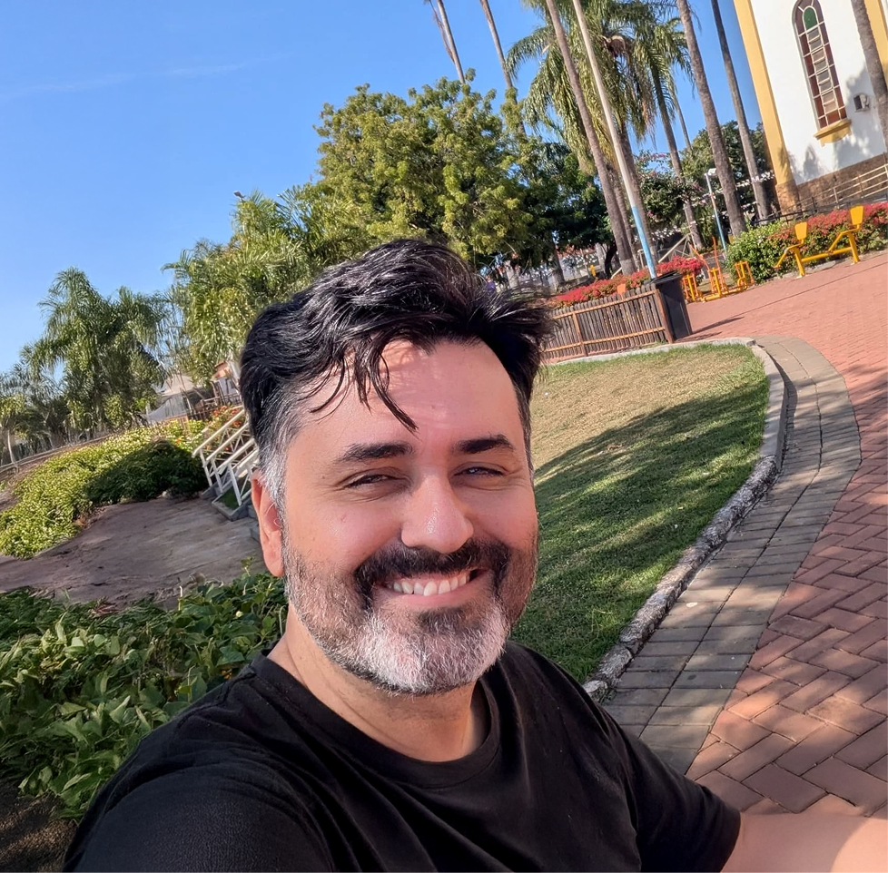 A man is taking a selfie outdoors, smiling, with a park that has green trees, palm trees, a brick pathway, yellow benches, and a white building with arched windows in the background.