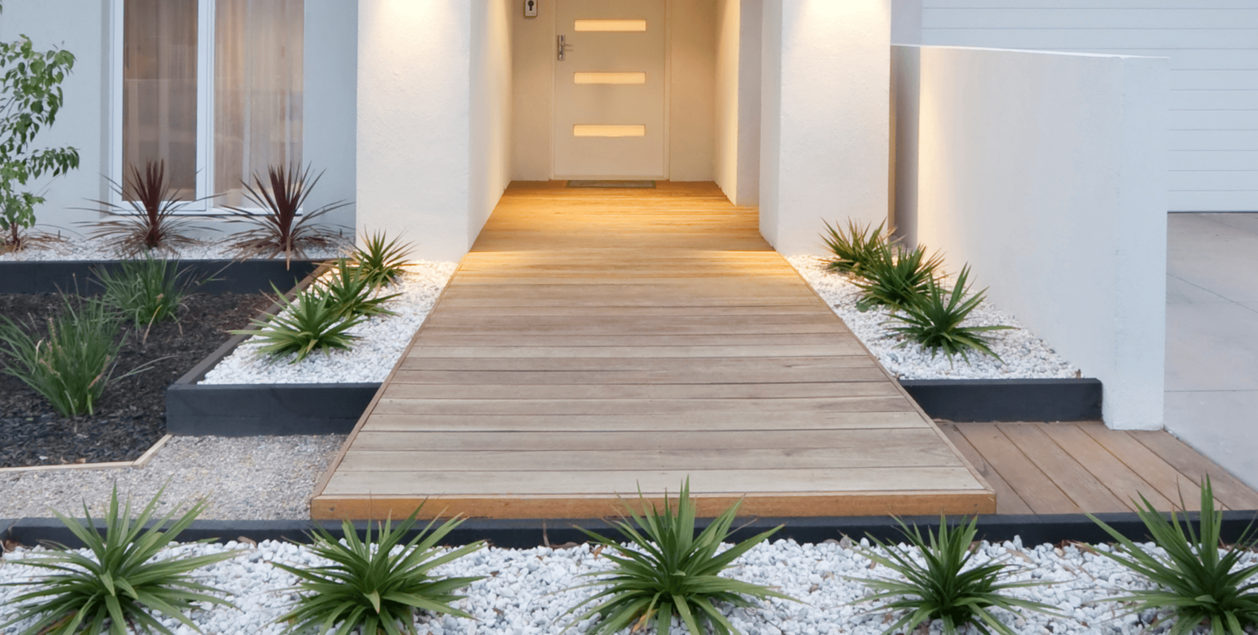 Front porch with wooden walkway, landscaped with green and purple plants on white gravel, leading to a door with horizontal windows, white walls, and soft lighting.