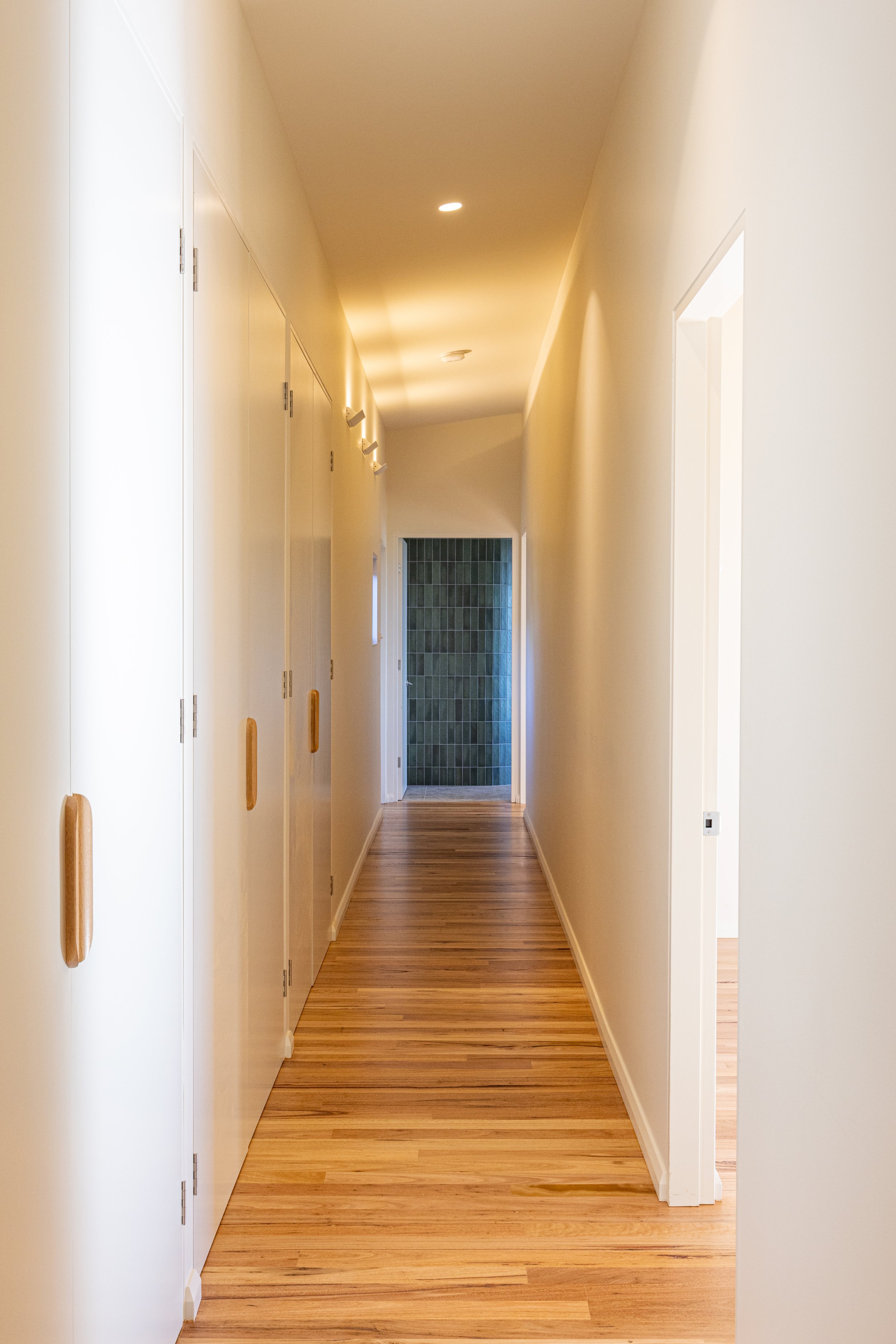 A hallway with wooden flooring, cream-colored walls, white doors with wooden handles on the left, and a doorway at the end revealing a room with dark green tiles.