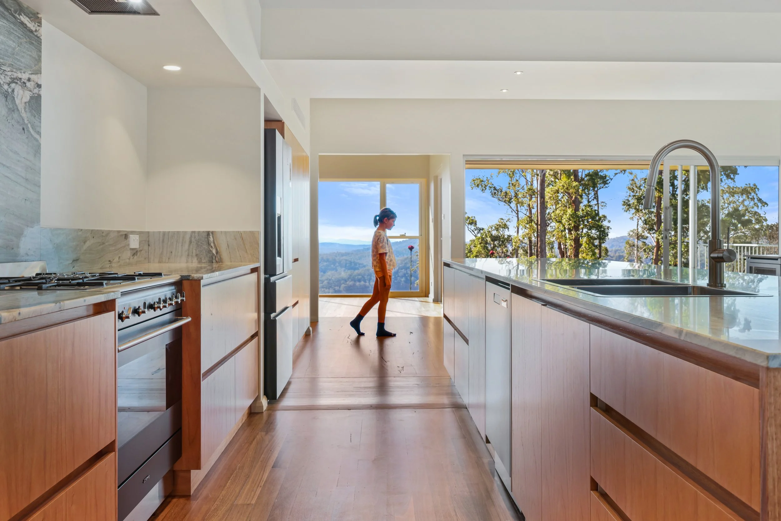 A woman standing in a modern kitchen with large windows showing a view of trees and mountains, white cabinets, marble backsplash, and stainless steel appliances.