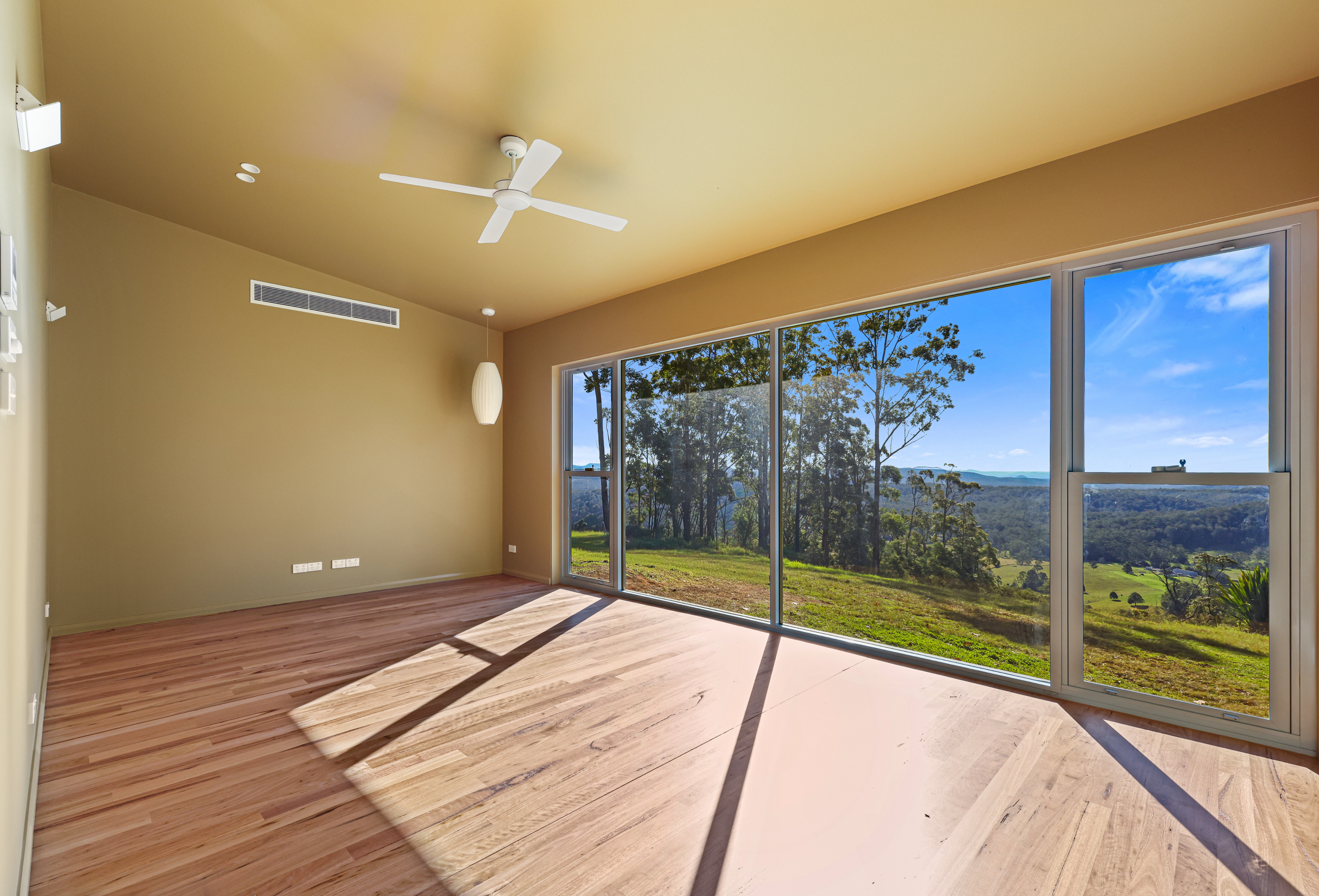 Empty room with large glass windows showing a scenic outdoor landscape of trees and hills, wood flooring, a ceiling fan, and a hanging light fixture.