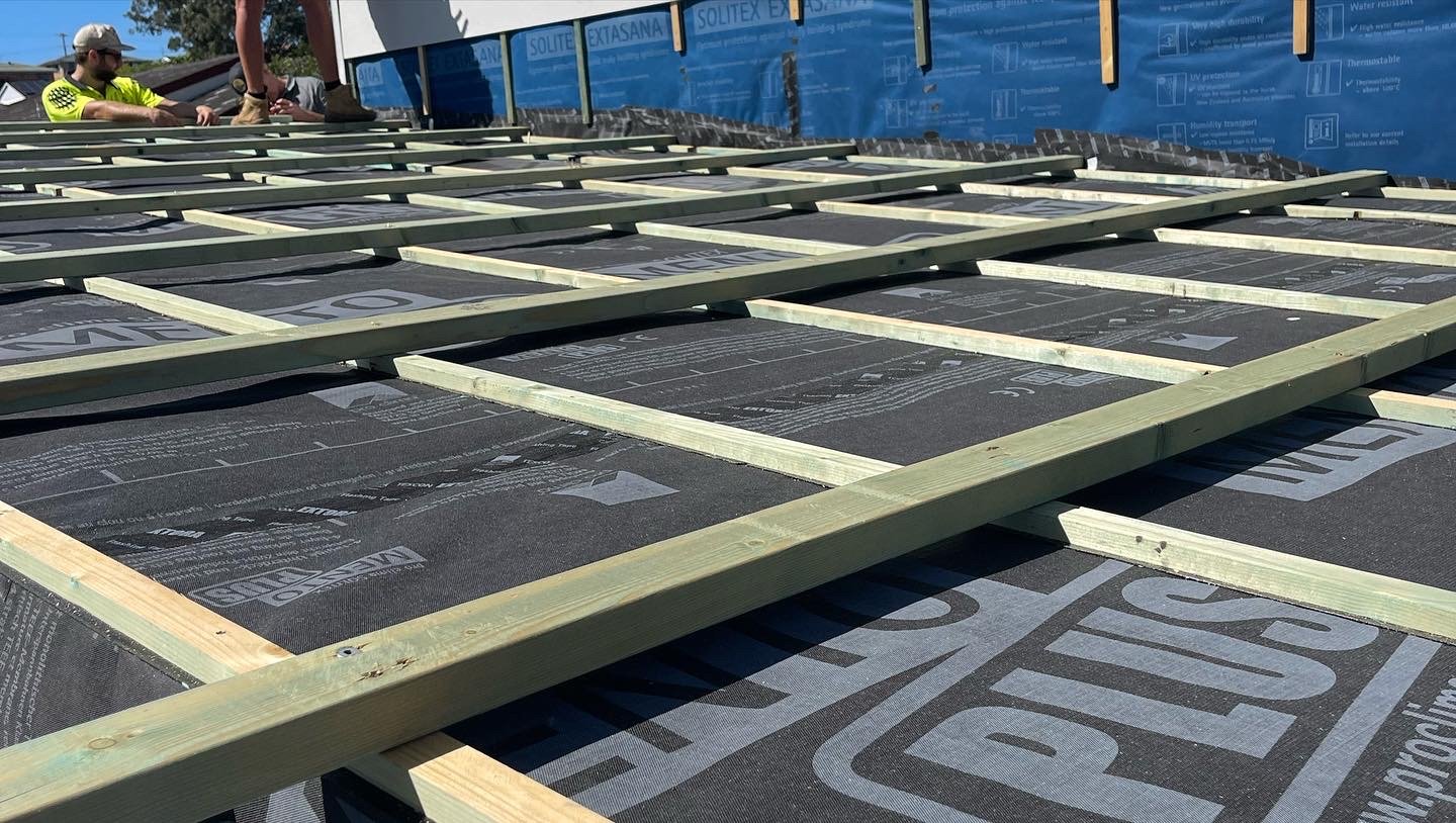 Construction workers installing roofing materials on a house, with wooden framing and black weatherproof underlayment visible.