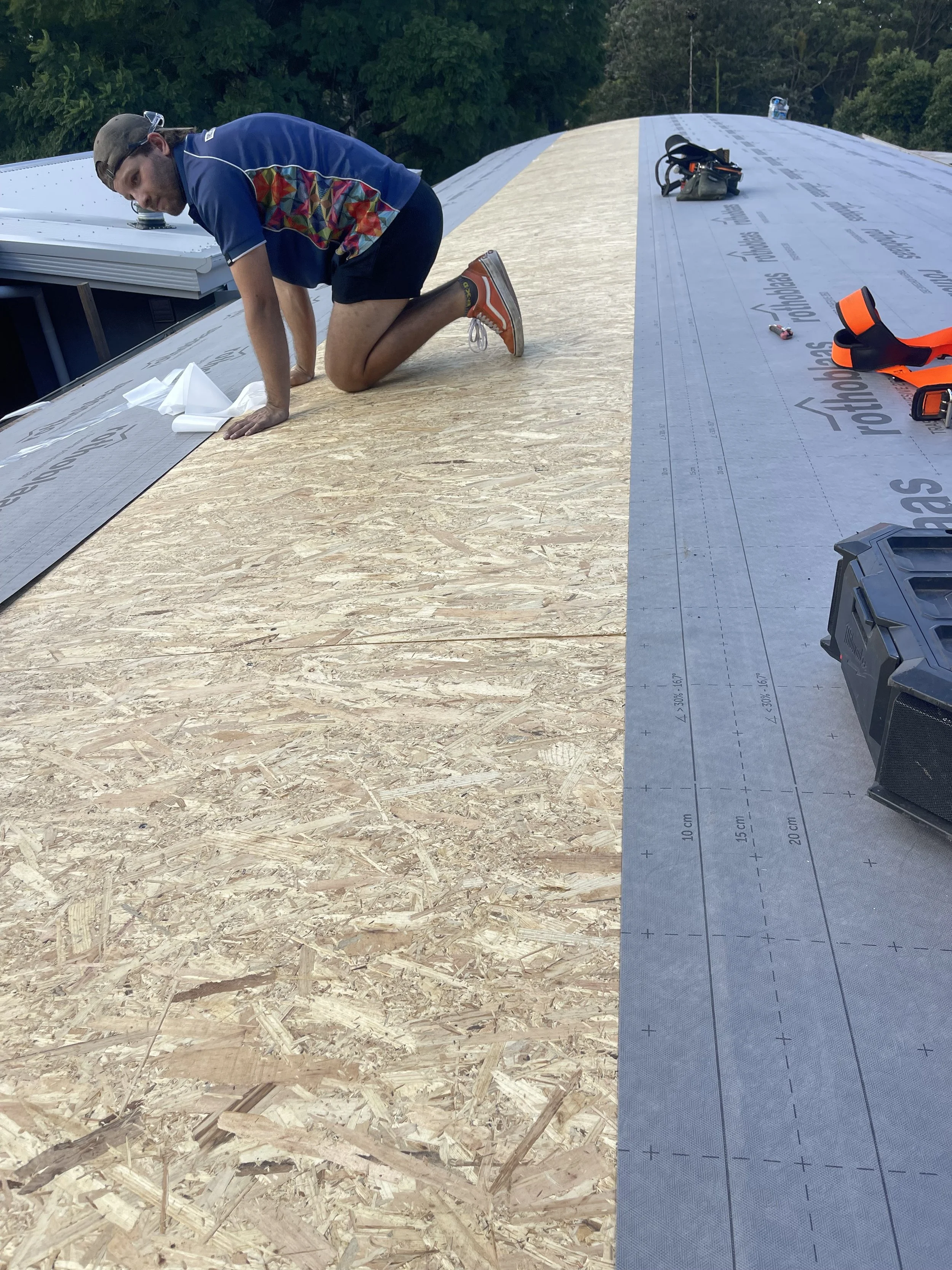 A man working on an unfinished roof, kneeling on a wooden surface with building tools and equipment nearby, with trees in the background.