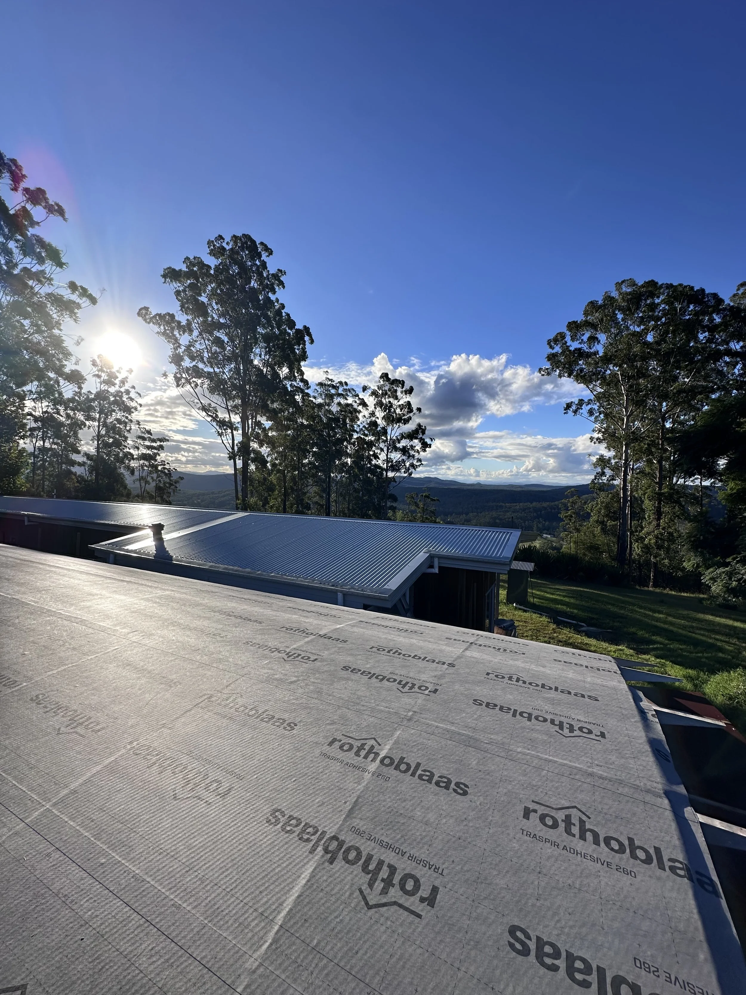 A view of a rooftop under construction, covered with roofing underlayment, with trees and a blue sky with some clouds in the background.