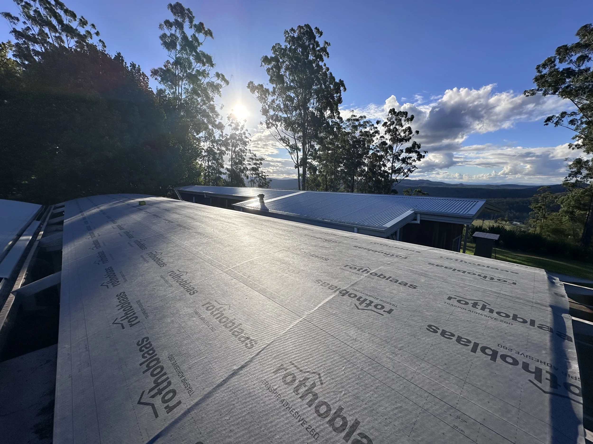 View from near a building with a metal roof, overlooking a landscape with trees and distant hills, under a partly cloudy sky with the sun shining through the trees.