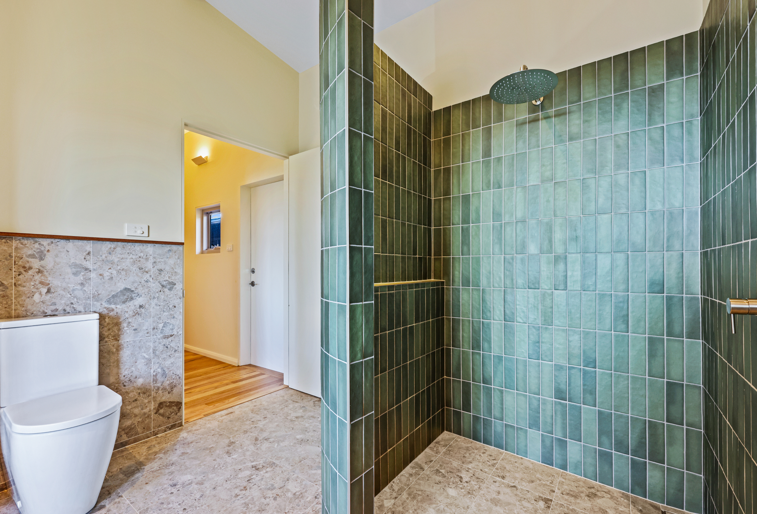 A modern bathroom featuring a green-tiled walk-in shower with a rainfall showerhead, a white toilet, and a partial view of a yellow wall with a small window and a hallway with a wooden floor.