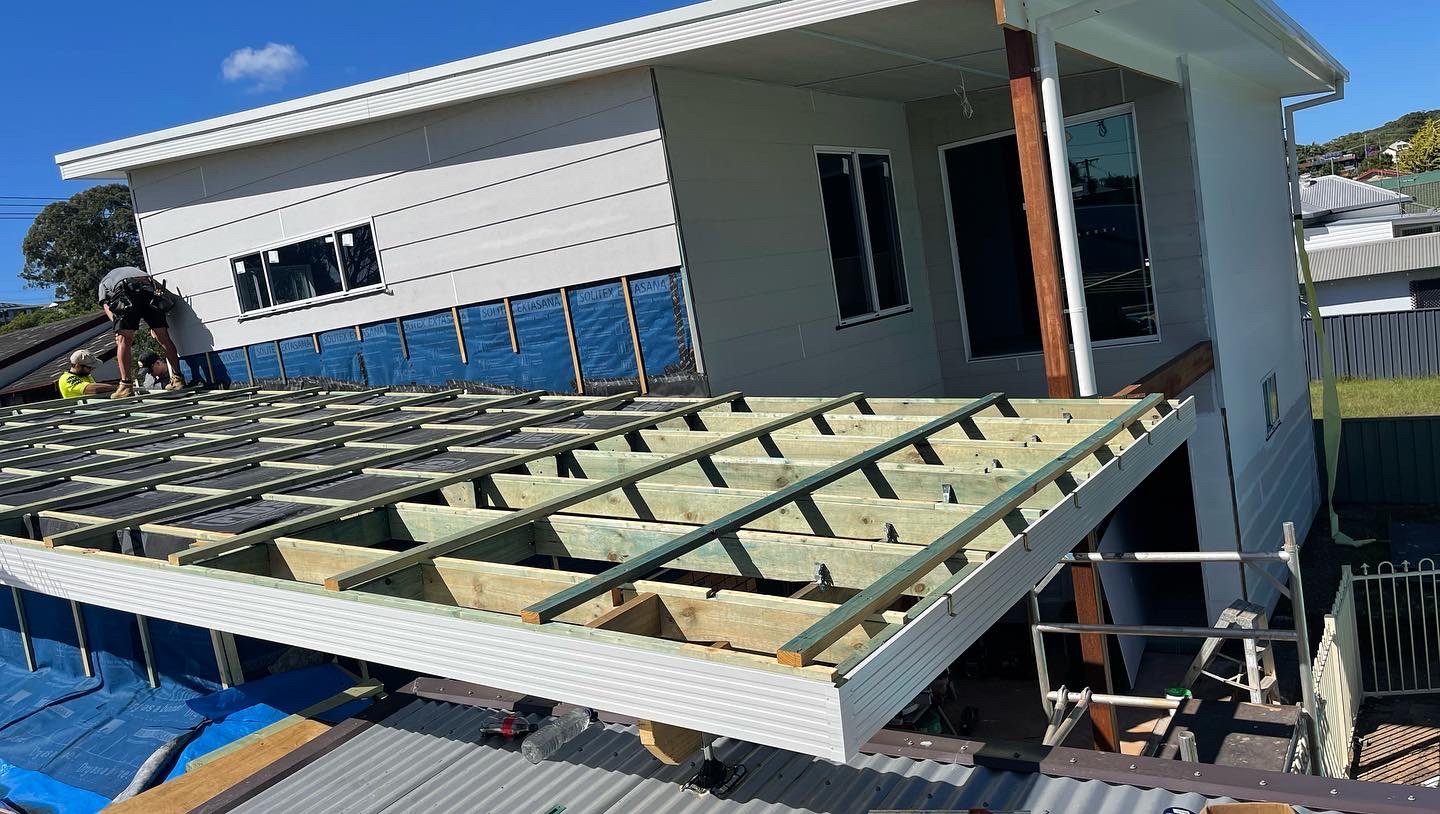 Construction workers installing a roof on a house with an extending section. The house is partially finished, with some siding installed and other areas still under construction.