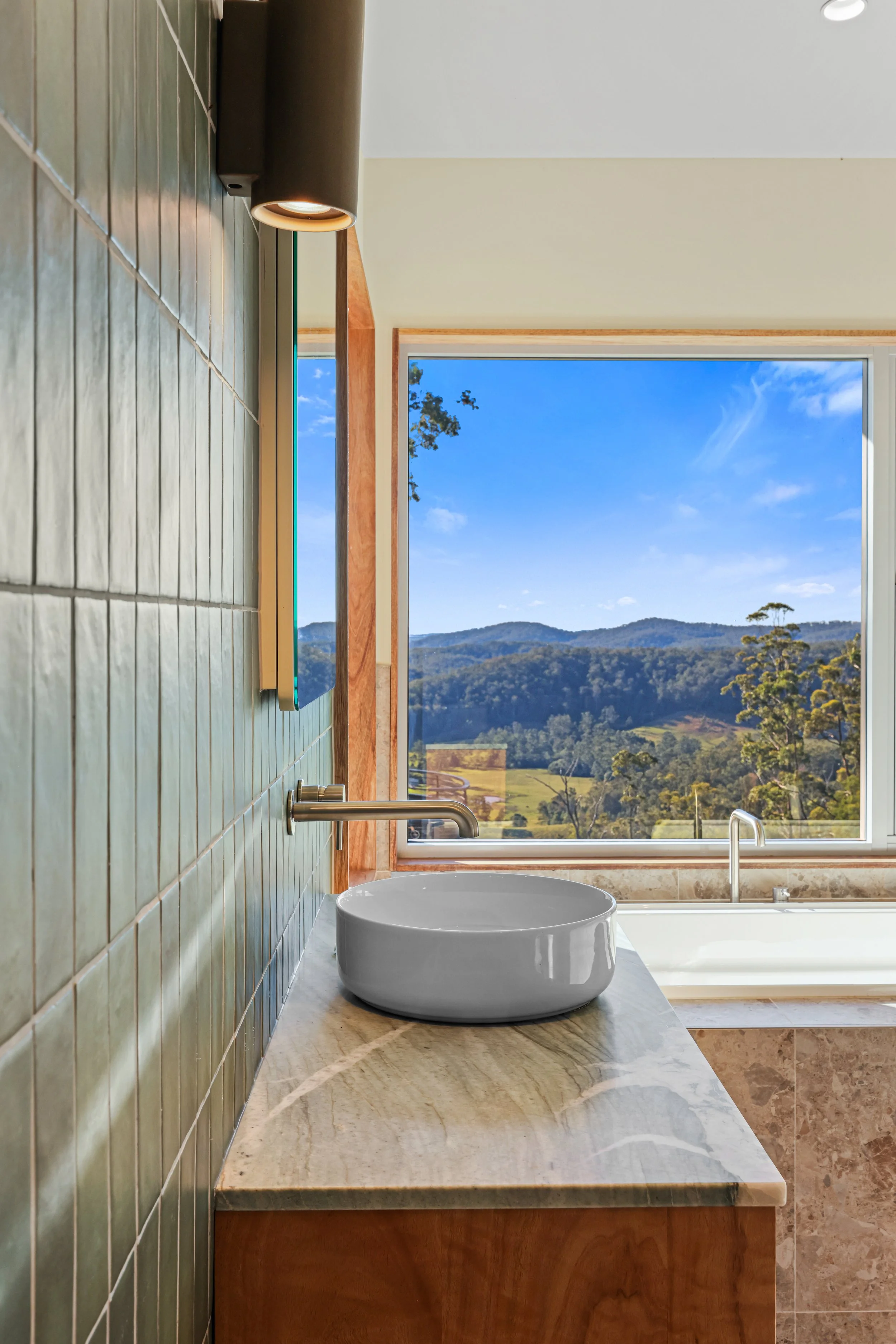 Bathroom with a round white basin sink on a marble countertop, green tiled wall, large window with mountain view, wooden trim, and a bathtub.