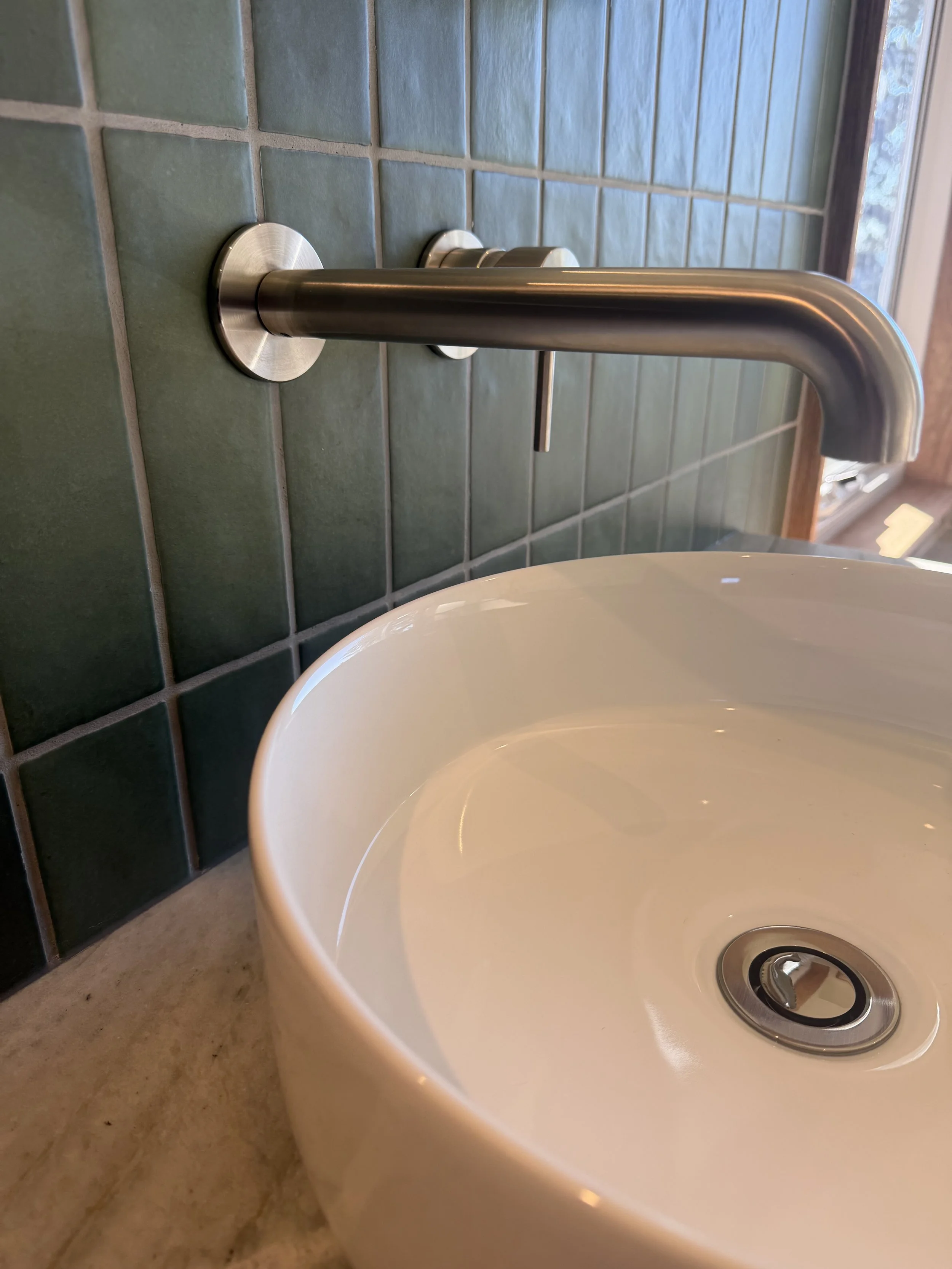 Close-up of a white ceramic bathroom sink with metal drain, next to teal tiled wall, and part of a countertop.
