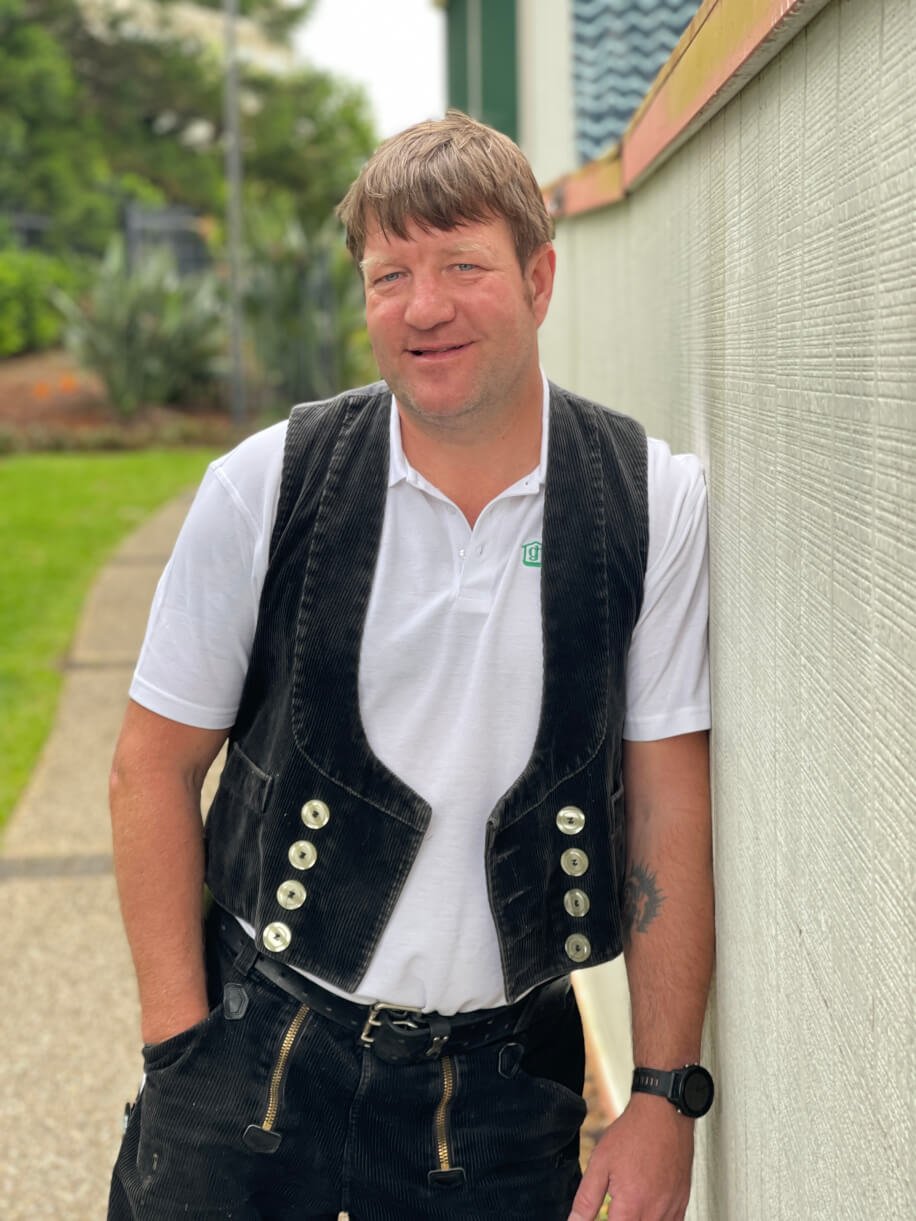 A man with light brown hair and a slight smile, wearing a white Polo shirt, a black vest, and black pants with zippers. He has a tattoo on his lower arm and is standing outdoors leaning against a beige wall.