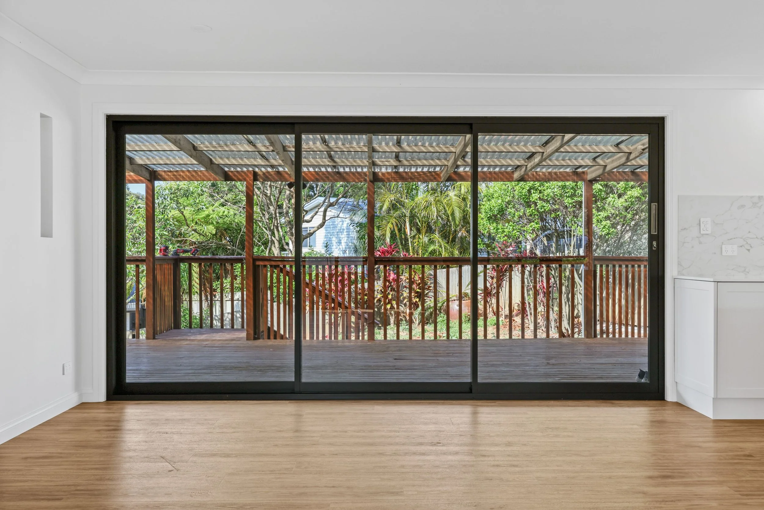 View through glass sliding doors onto a wooden deck with a railing, overlooking a lush yard with trees and plants under a metal roof canopy.