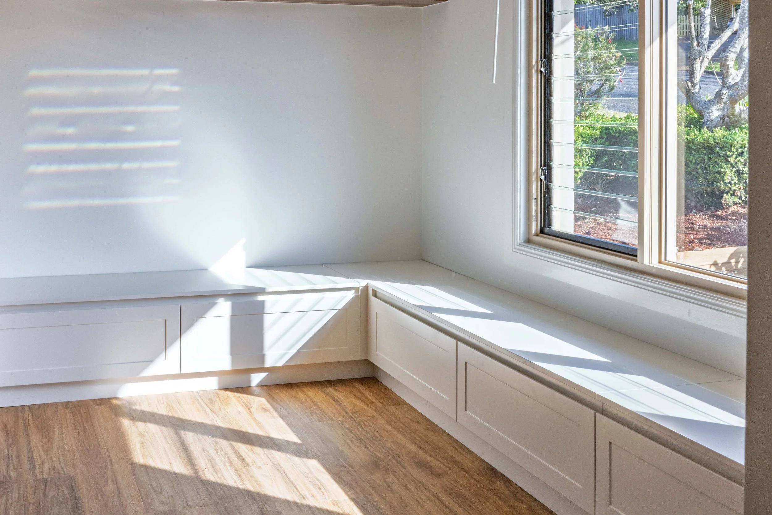 Sunlight streams through a window with blinds, casting shadows on a white wall and wooden floor in a bright room.
