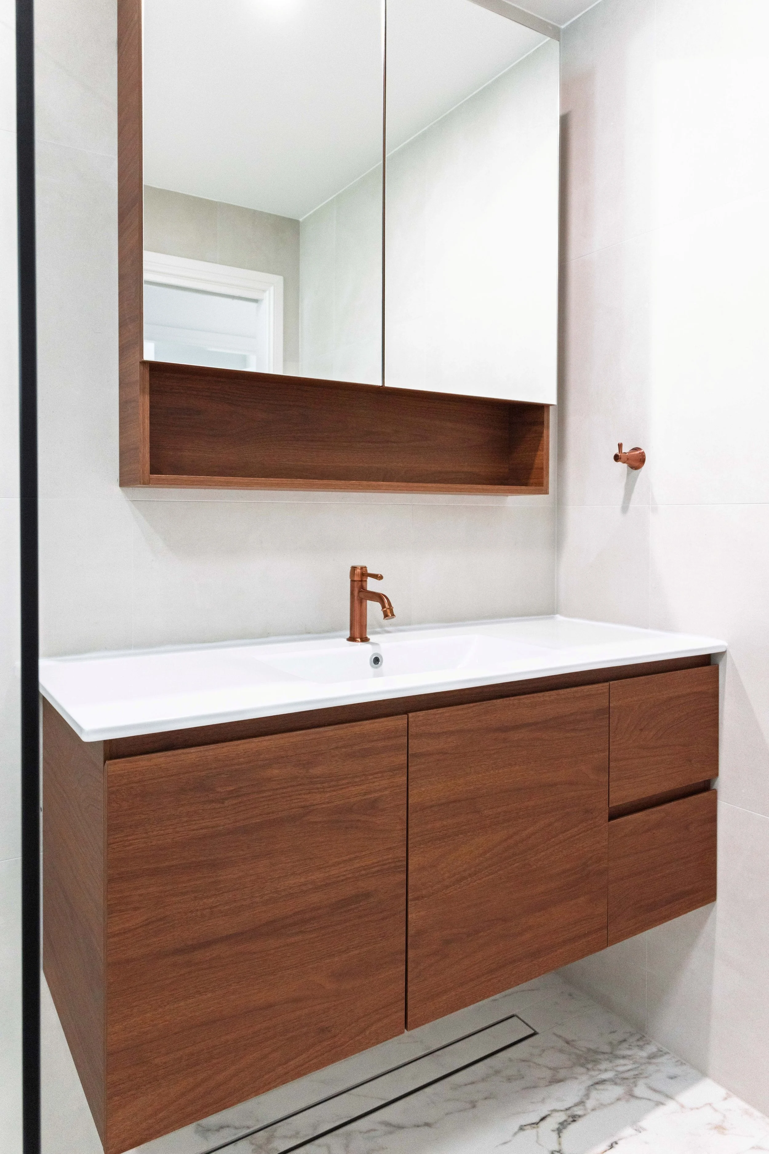 Modern bathroom vanity with a white countertop, a copper faucet, and a wooden cabinet with drawers. A large mirror is mounted above the vanity, and a small copper hook is attached to the wall on the right.