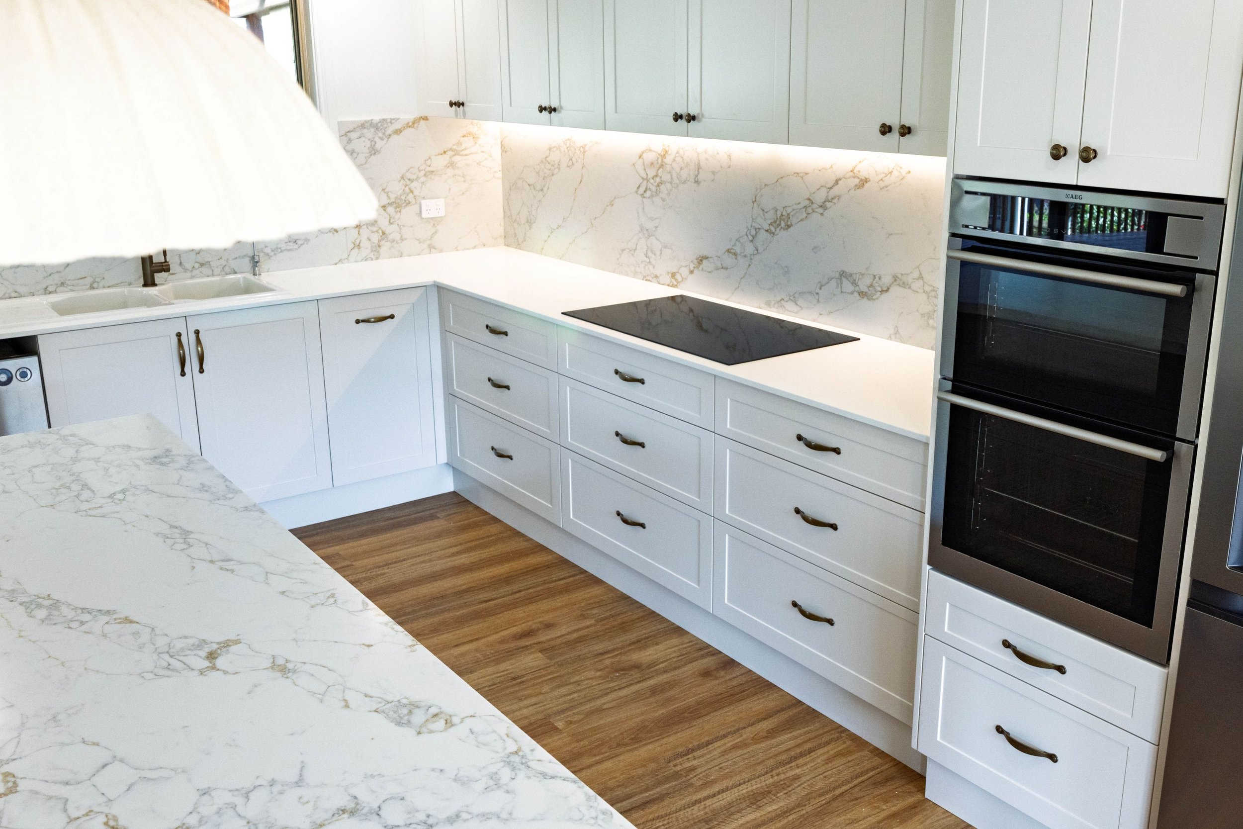 A modern kitchen with white cabinets, marble countertops, and a marble backsplash. The kitchen has a black electric cooktop, built-in oven, and a double sink. There is wood flooring and under-cabinet lighting.