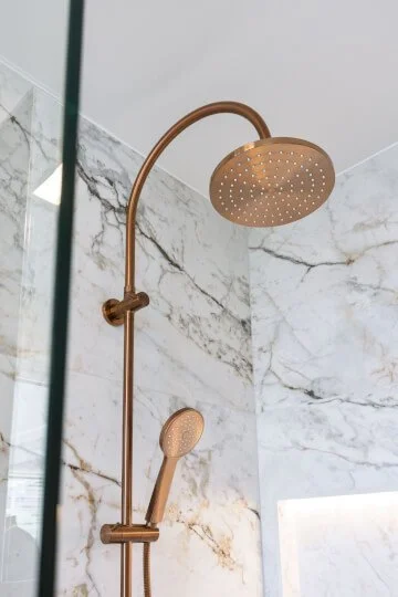 Touchless bronze shower head and handheld shower wand mounted on white marble wall in bathroom.
