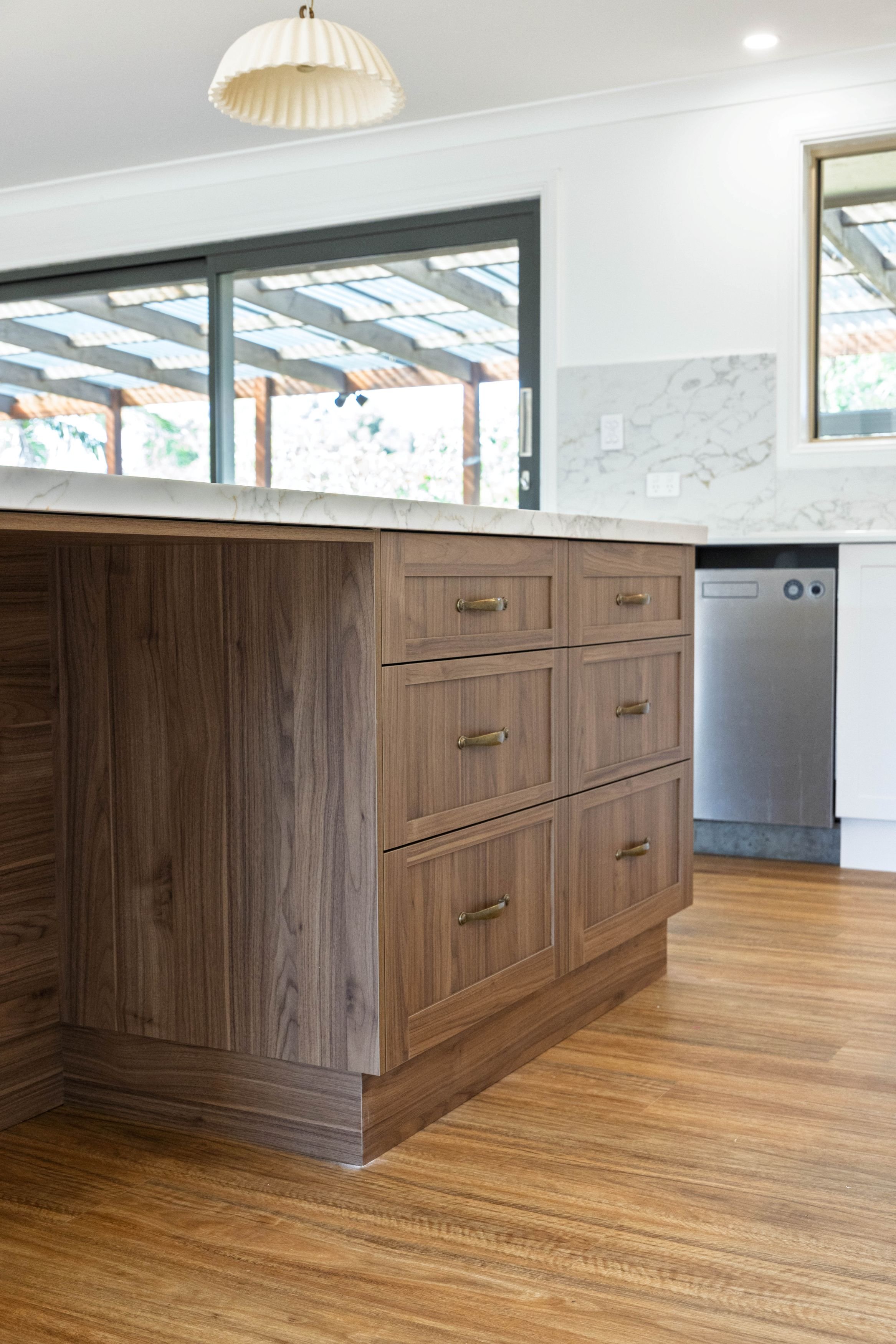 Close-up of a kitchen island with wooden drawers and a marble countertop, with a sliding glass door and window in the background. The kitchen has wooden flooring.