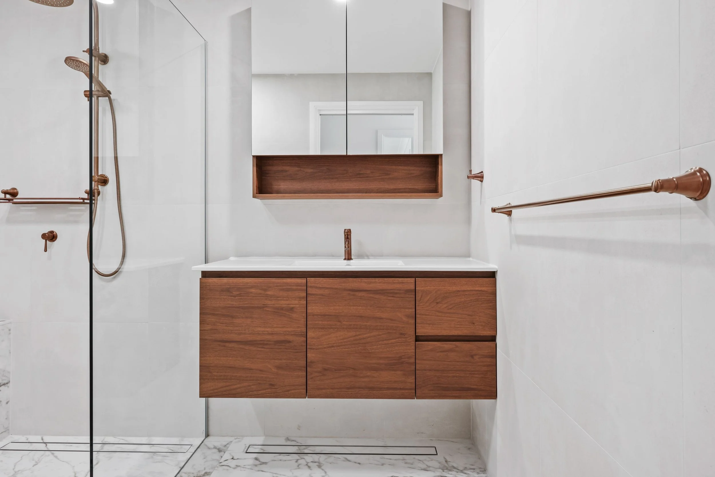 Modern bathroom with white walls, a wooden vanity with a white countertop, an oval mirror cabinet, copper fixtures, a walk-in shower with glass door, and marble flooring.