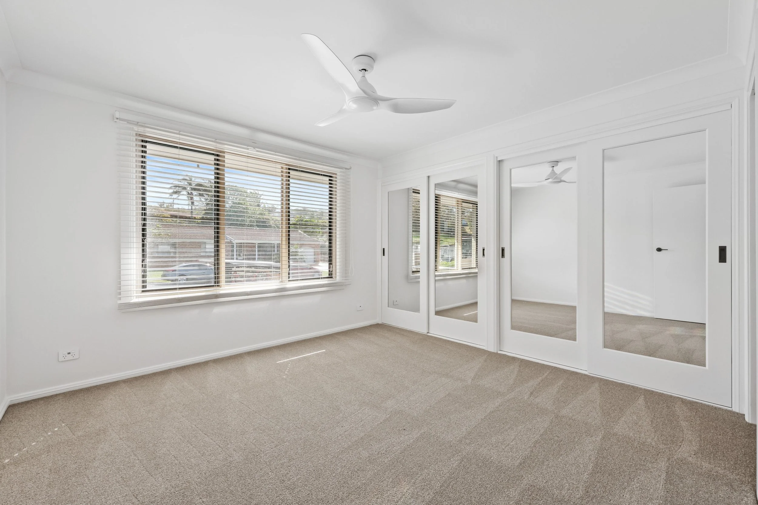 Empty bedroom with large window, white walls, mirrored closet doors, ceiling fan, and beige carpet.