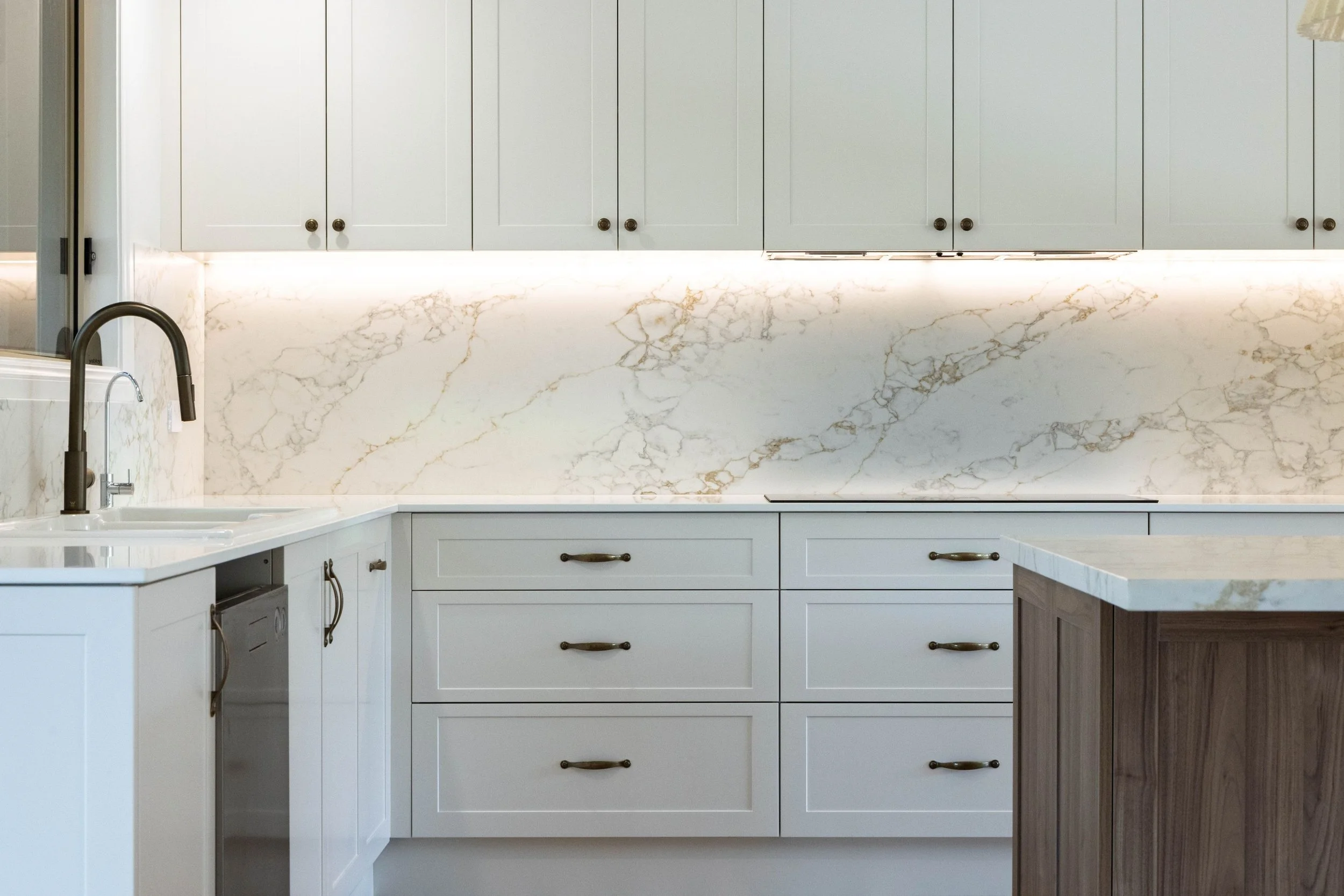 A kitchen with white cabinets, a marble backsplash, and a marble countertop. There is a black faucet and a sink on the left, and a brown and white kitchen island on the right.