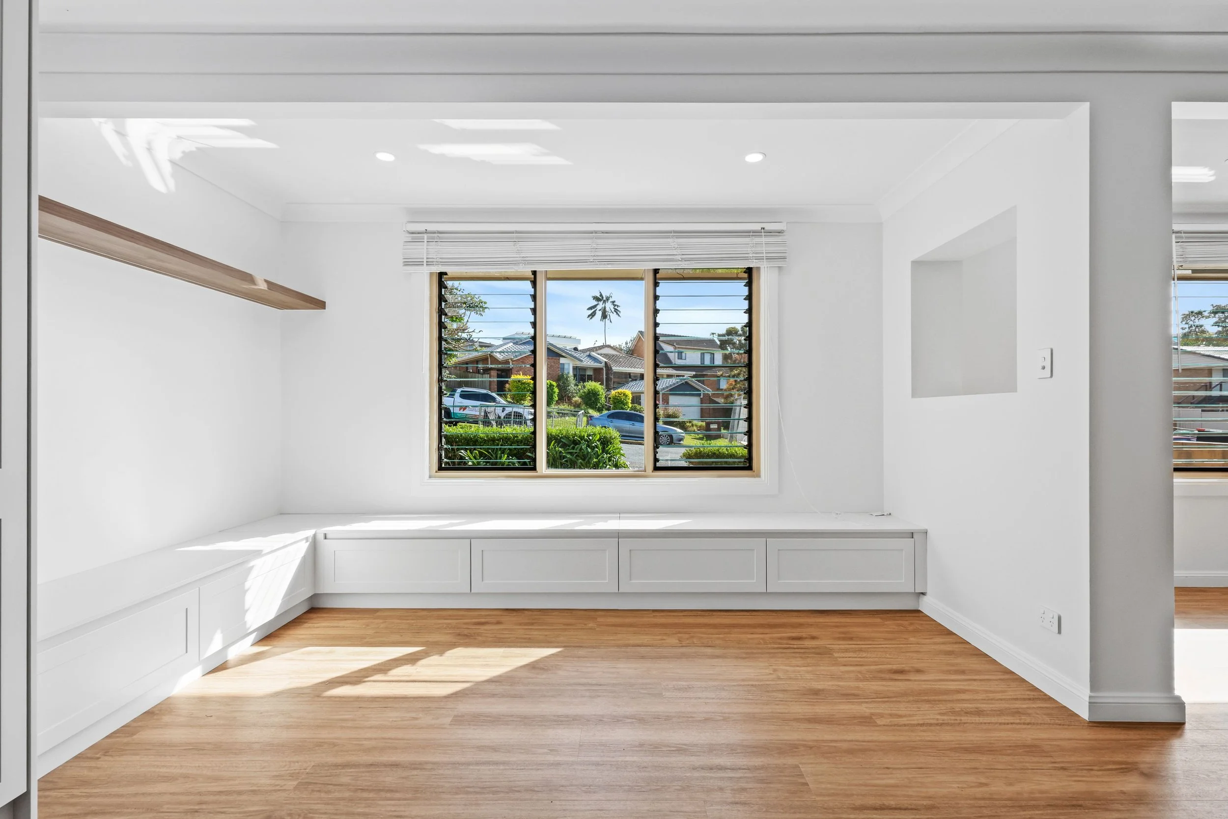 Empty white room with large window featuring horizontal blinds, built-in white bench, light wood flooring, and minimal decor.