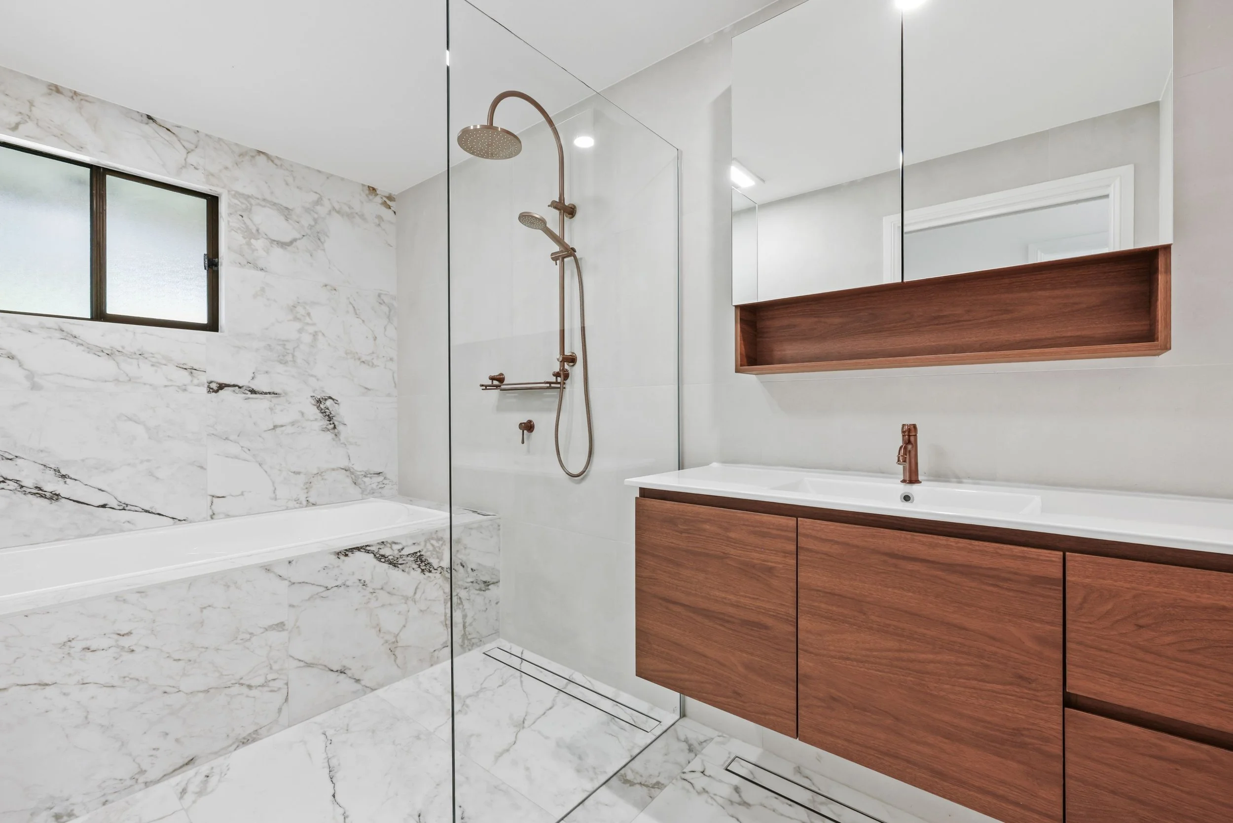Modern bathroom with a marble shower wall, a glass shower door, a copper showerhead, a wooden vanity with a white sink, a large mirror, and a frosted window.