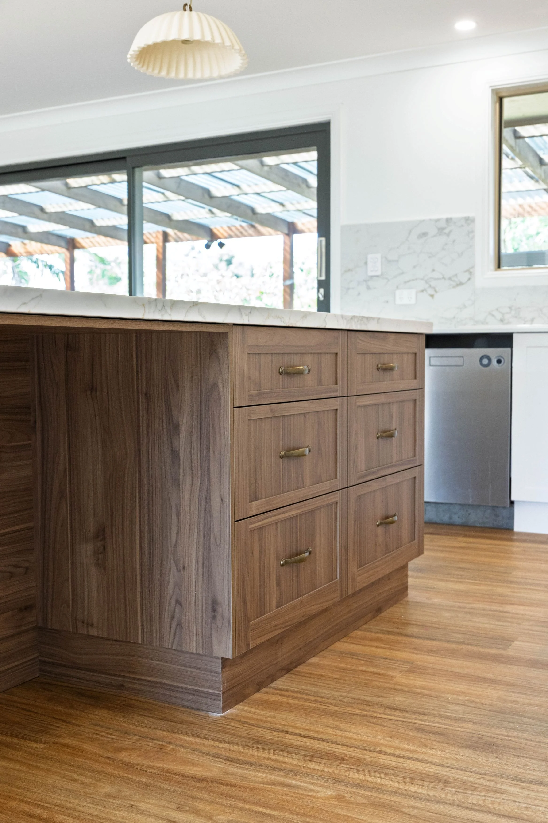 A kitchen island with wooden cabinets, a marble countertop, and brass handles. The floor is wooden, and there is a background view of an outdoor patio with a wooden pergola. The walls are white with a marble backsplash.