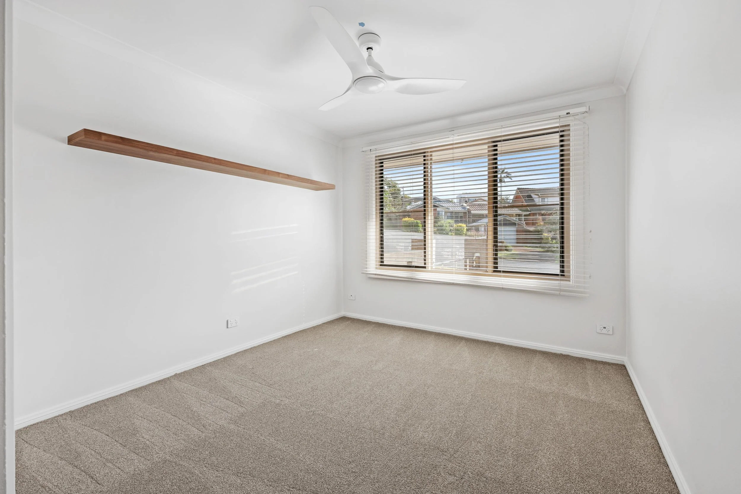 Empty room with white walls, beige carpet, a wooden shelf on the left wall, a large window with blinds, and a white ceiling fan.