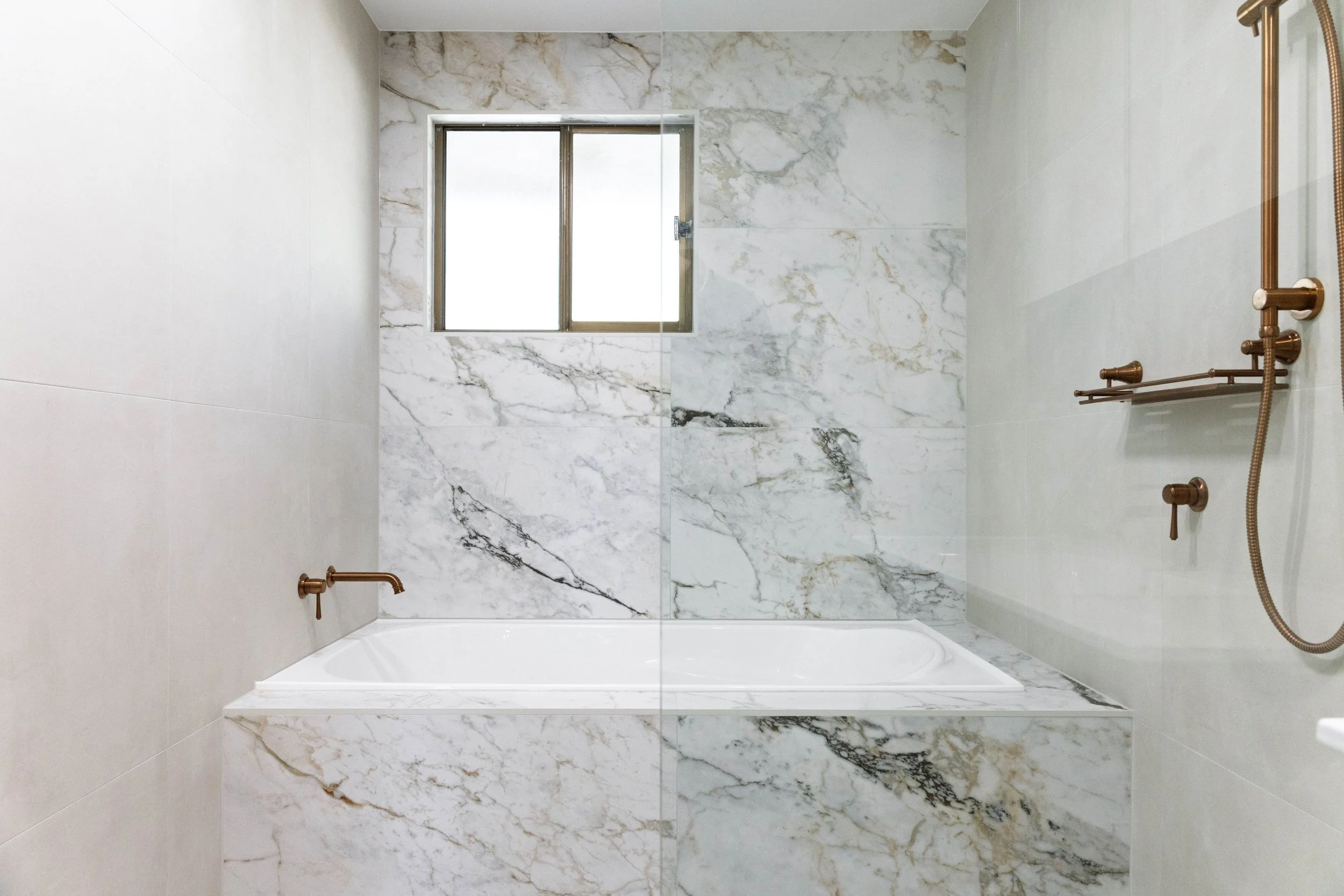 Modern bathroom with marble walls, a window, bronze fixtures, and a showerhead.