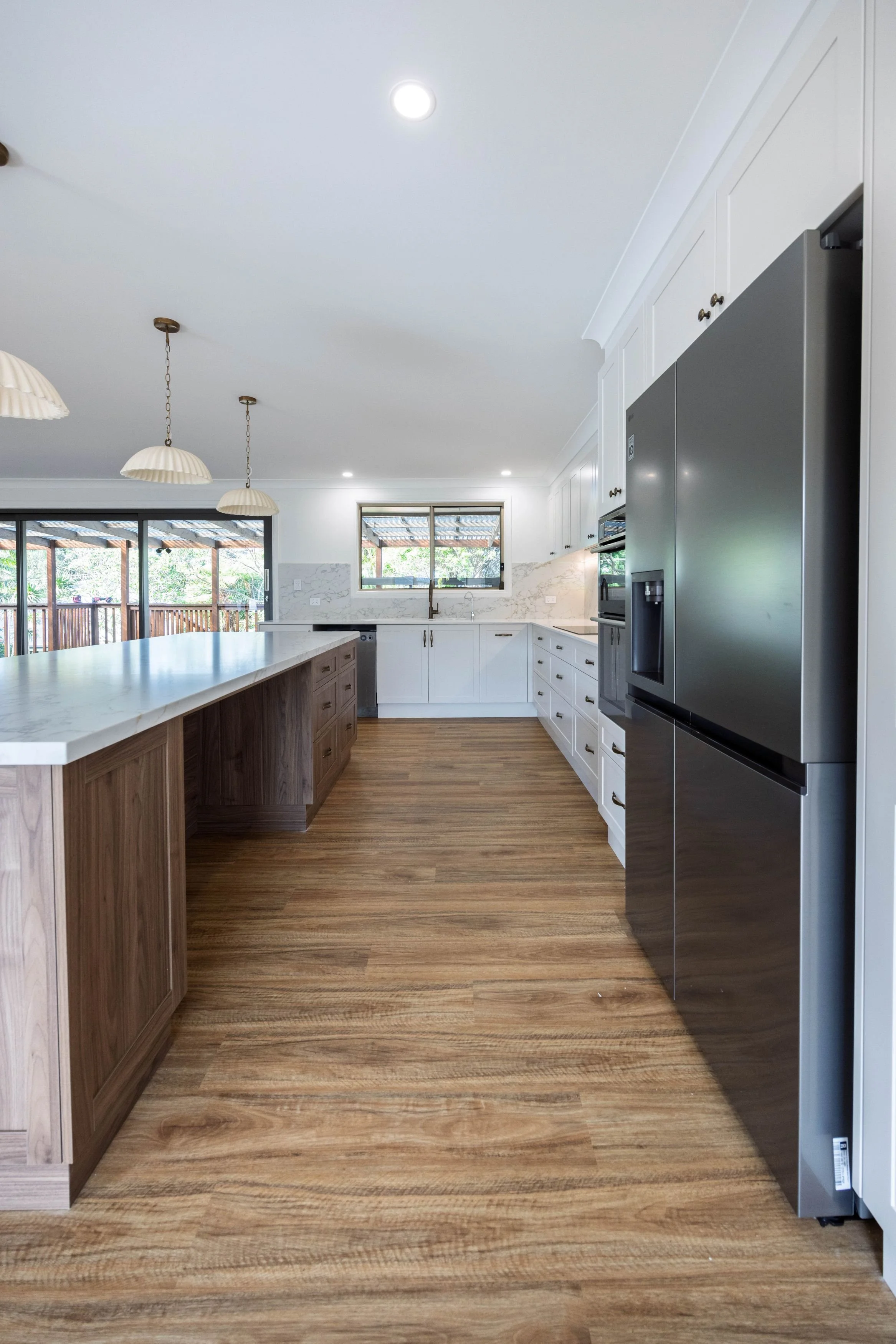 Modern kitchen with white cabinets, marble countertops, a stainless steel refrigerator, hardwood flooring, and pendant lights hanging above the island. Large windows provide natural light.