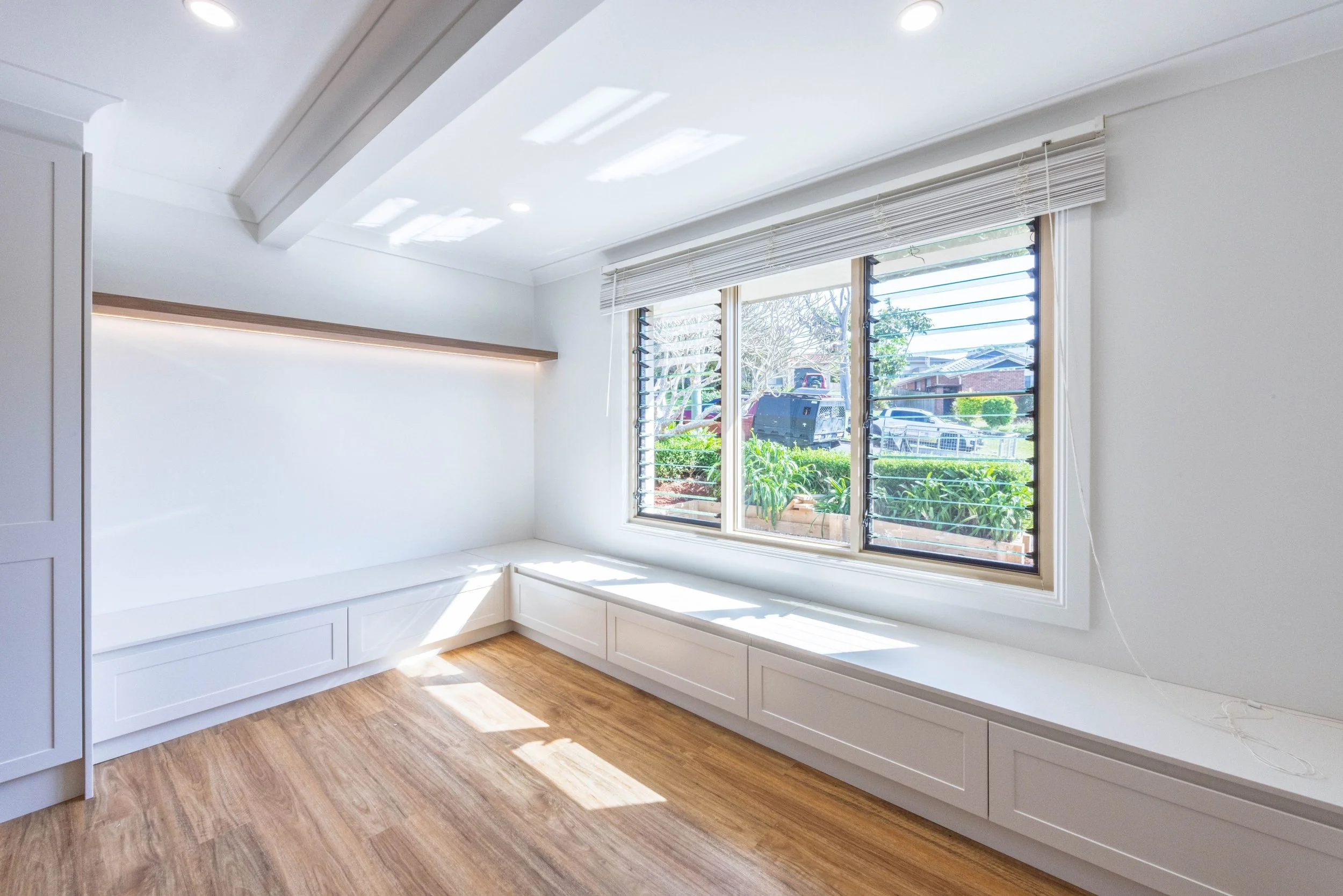 Bright room with large window, white window blinds, built-in white bench with storage, wooden flooring, and ceiling lights.