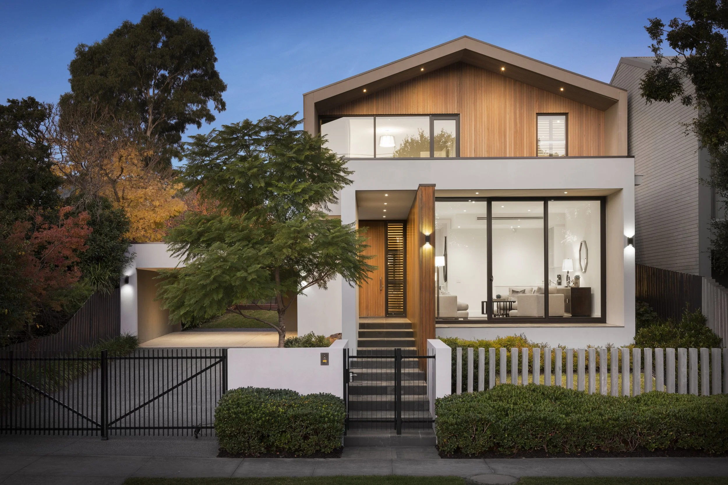 Modern two-story house with large glass windows, wooden accents, surrounded by trees and a black metal fence at dusk.