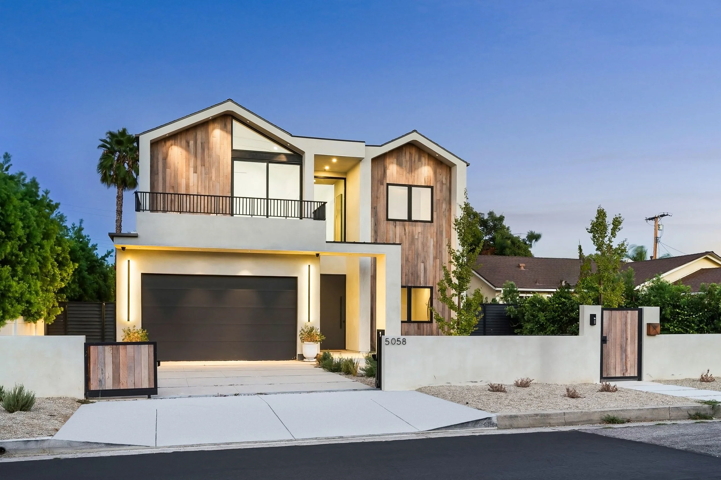 Modern two-story house with a white and wood exterior, black garage door, and front yard with plants and trees, located at 5058, in a suburban neighborhood during dusk.