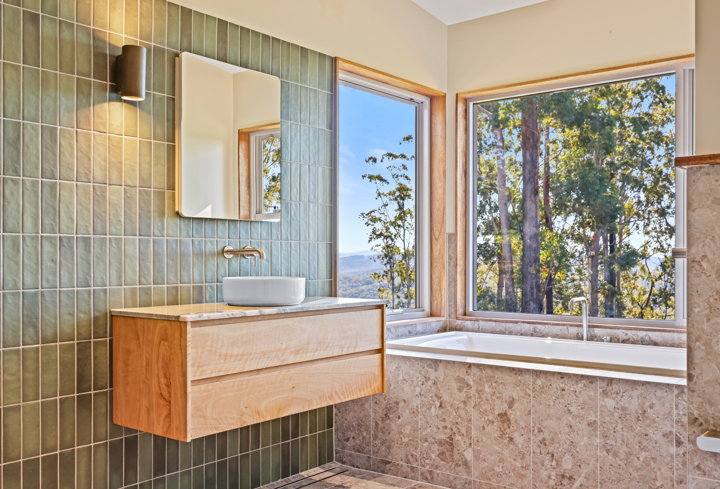 A bathroom with green tiled wall, a vanity with a white vessel sink, a wooden cabinet below, large windows showing trees and blue sky, and a bathtub beneath the windows.
