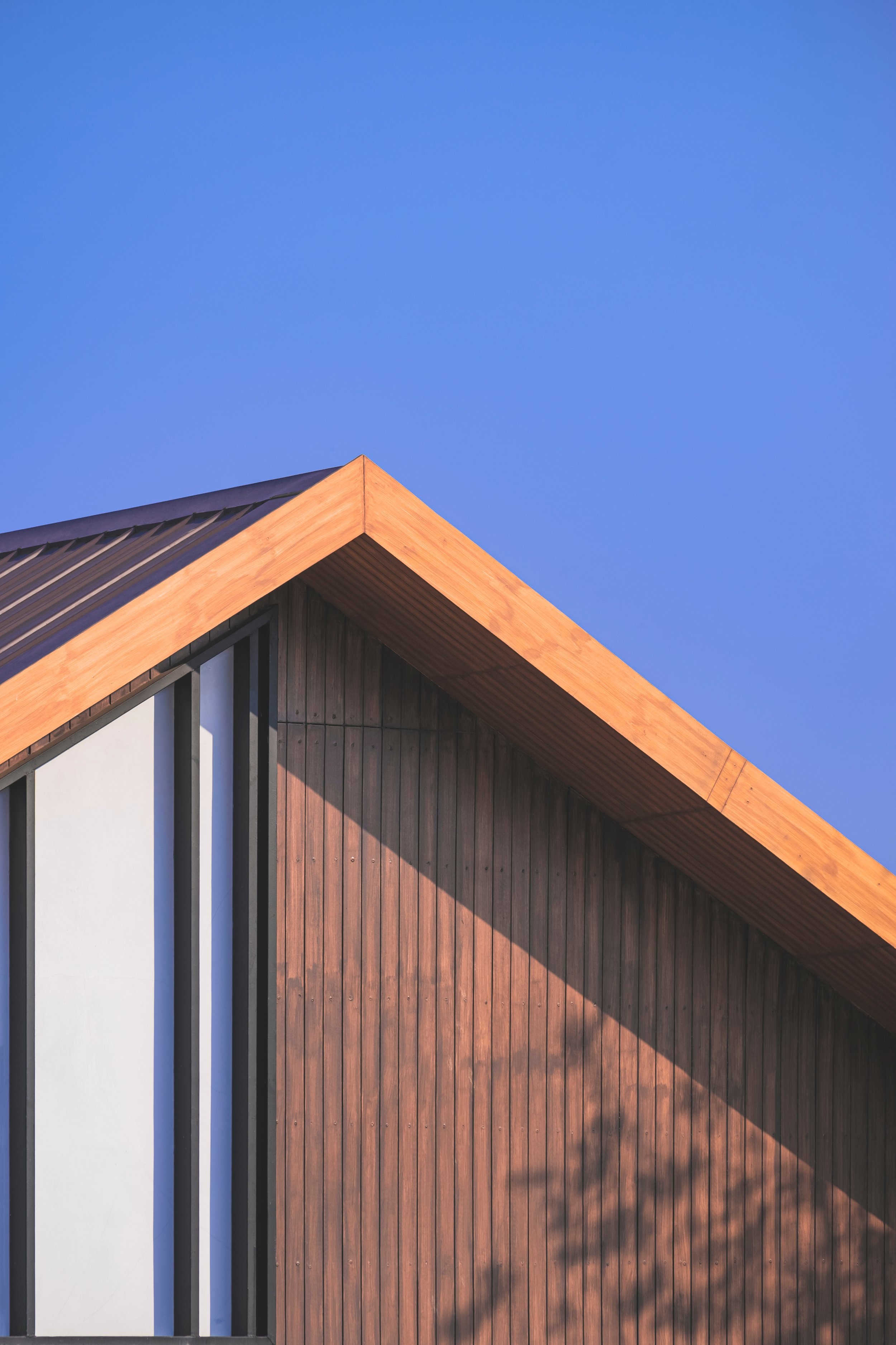 The image shows the corner of a modern building with a wooden roof and wall, black vertical and horizontal accents, and a clear blue sky in the background.