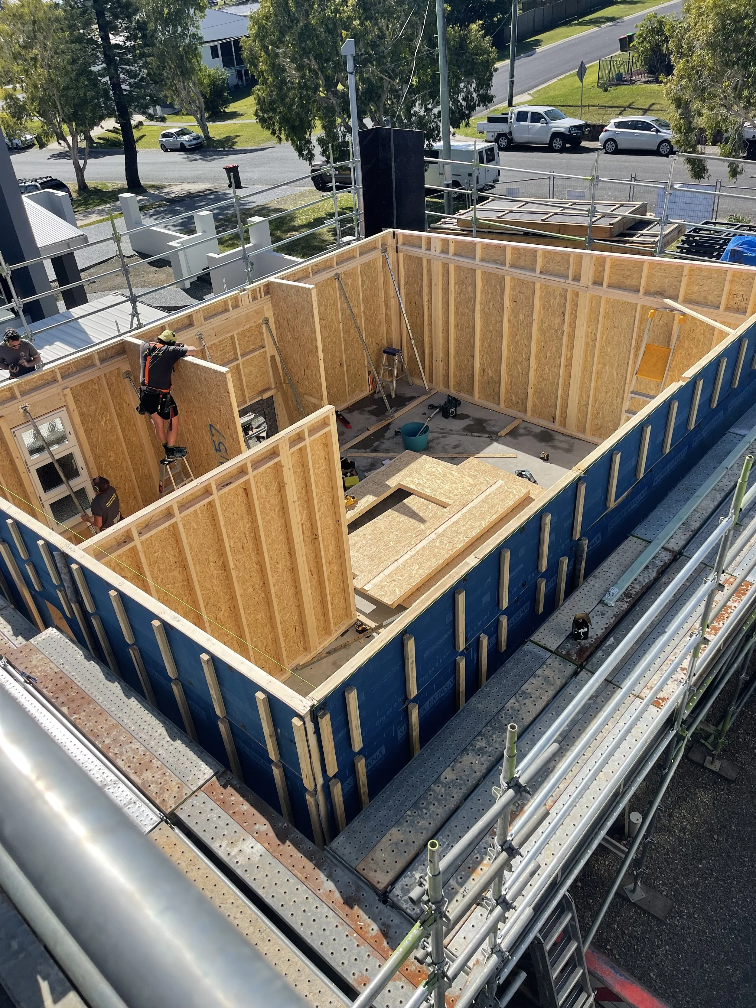 Construction workers building wooden walls for a house, with scaffolding surrounding the structure on a sunny day.