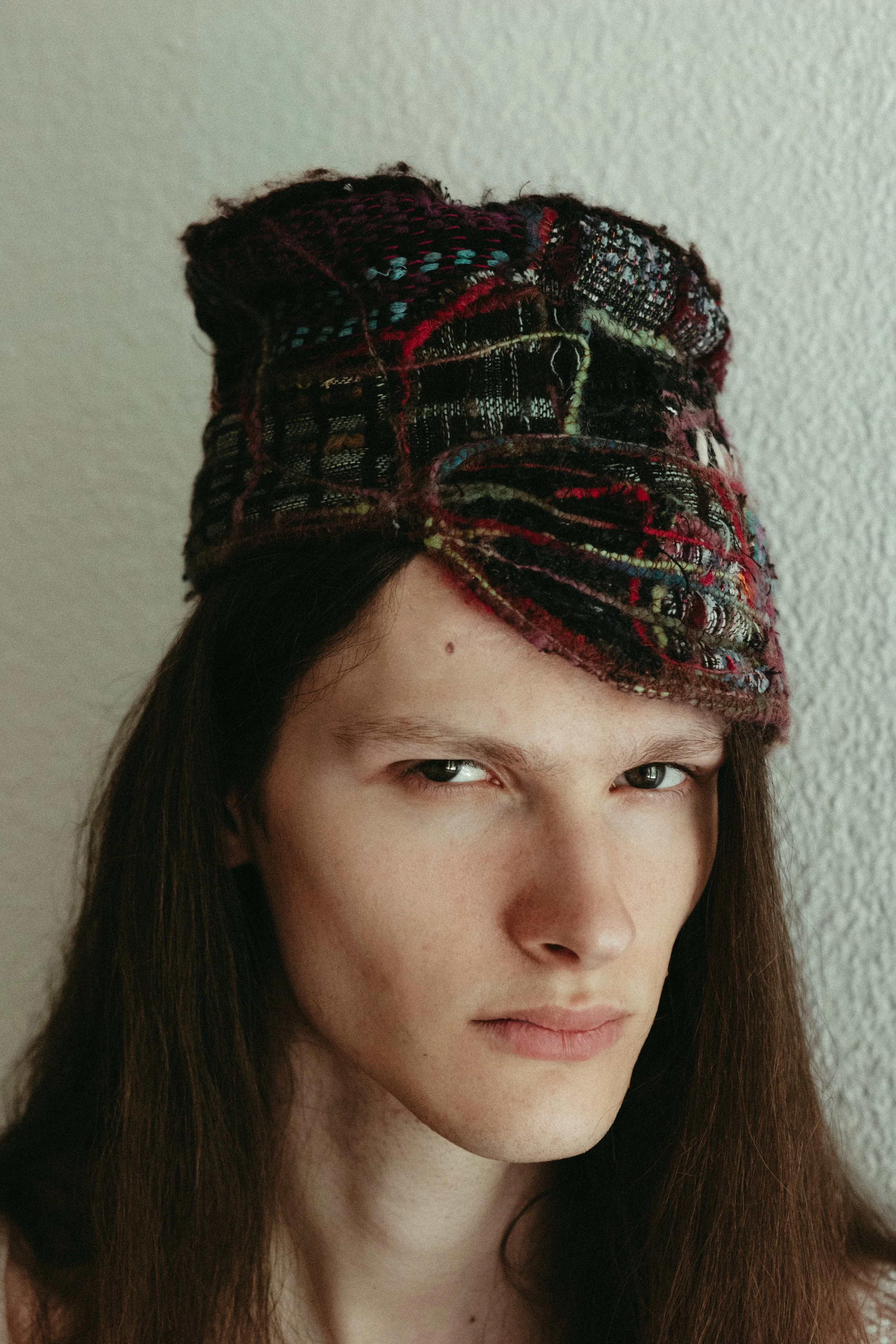 A woman with long brown hair wearing a colorful, textured, patterned headwrap or hat, standing against a plain, light-colored wall.