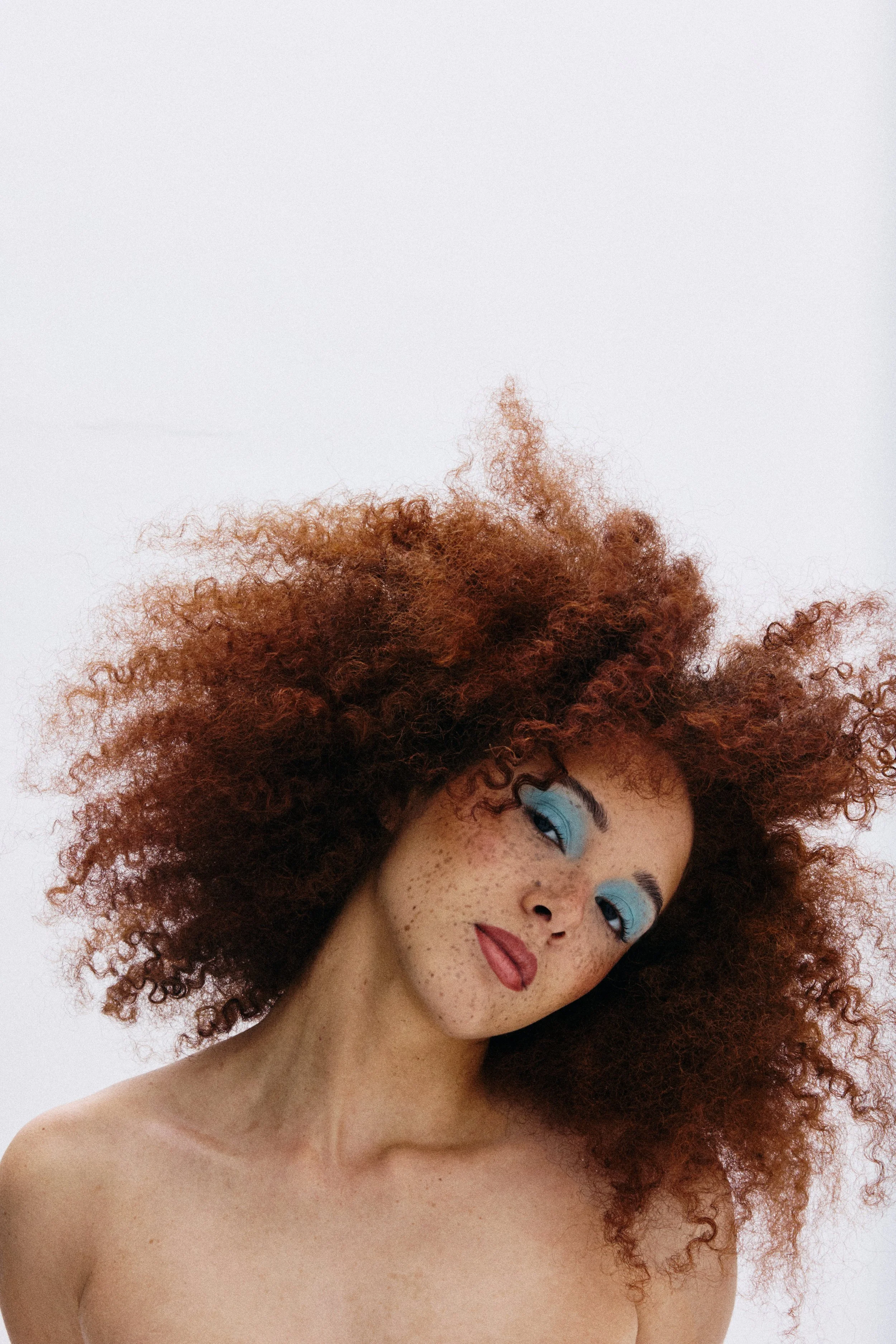 A woman with curly red hair, blue eyeshadow, freckles, and red lipstick, looking at the camera with a slight tilt of her head.