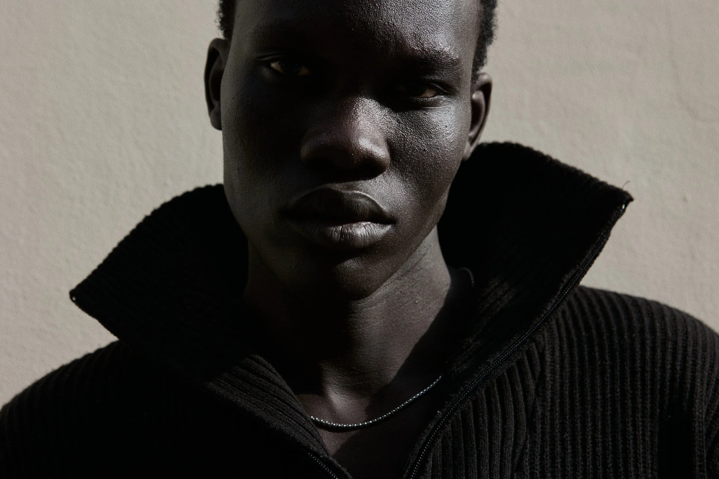 A close-up portrait of a person with dark skin, short black hair, wearing a black zip-up sweater with a textured knit pattern, against a plain light-colored background.