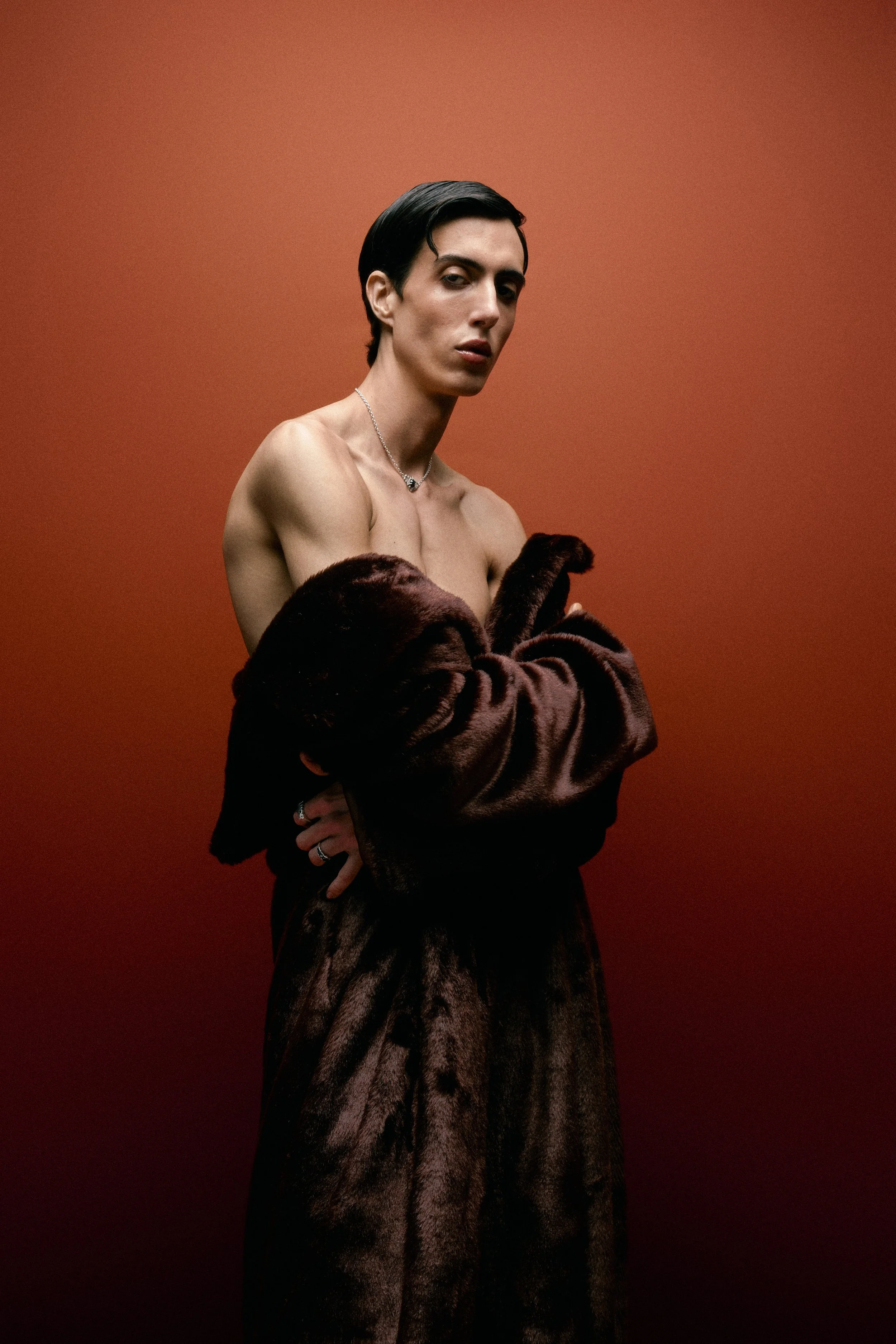 A person with short black hair, wearing a fur coat draped over their shoulders, stands against a reddish-brown background. They are shirtless, wearing a silver necklace, with a confident expression and arms crossed.