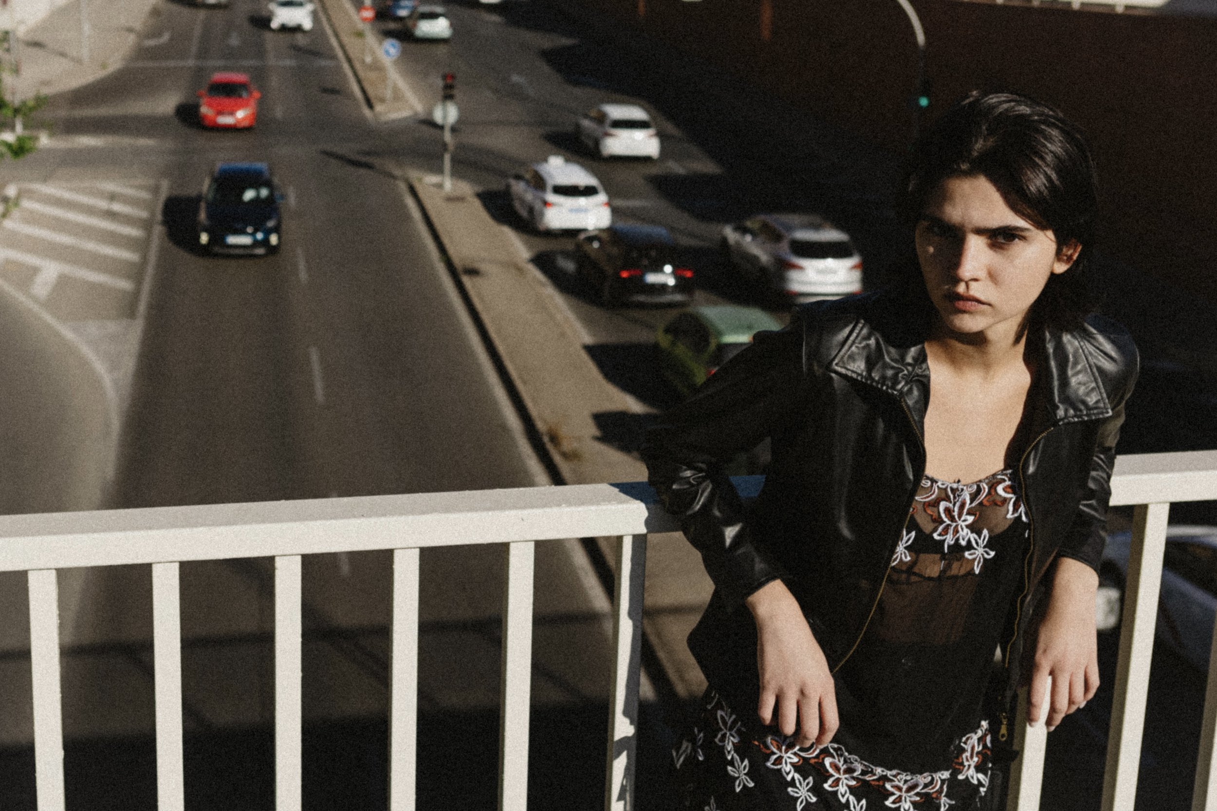 A woman with short dark hair, wearing a black leather jacket and a floral dress, leaning on a white railing on a sidewalk, with a parking lot filled with cars in the background during nighttime.
