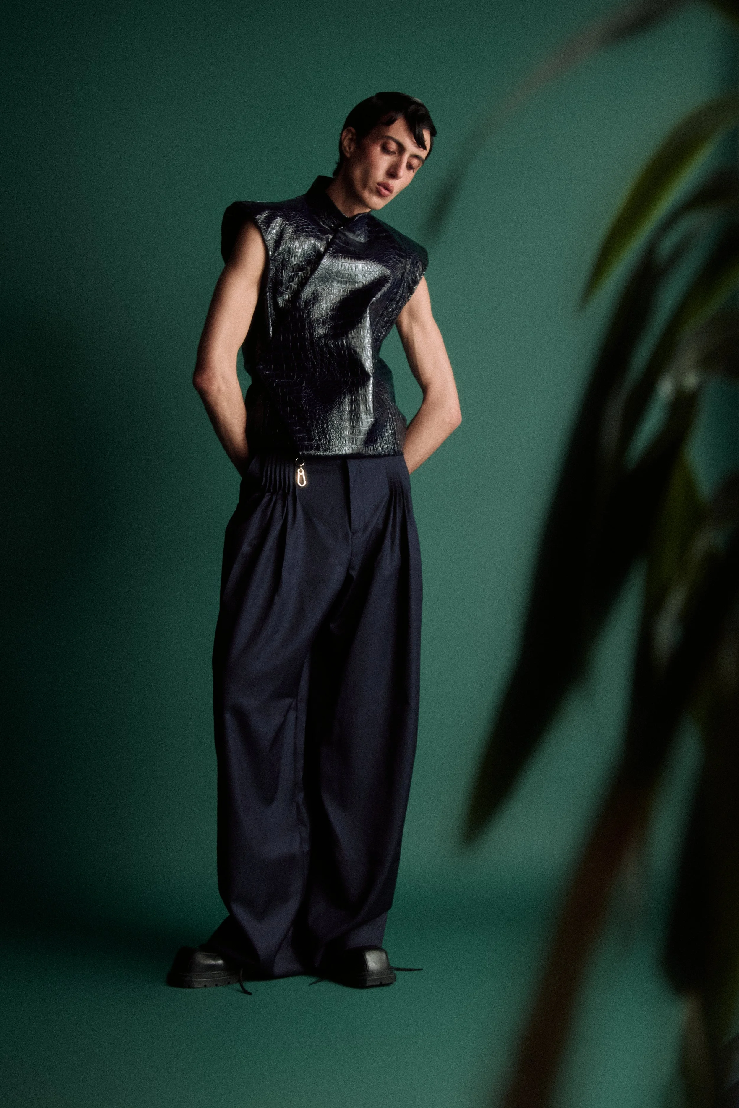 A woman with short dark hair wearing a shiny black sleeveless top and loose dark pants stands against a green background, looking downward.