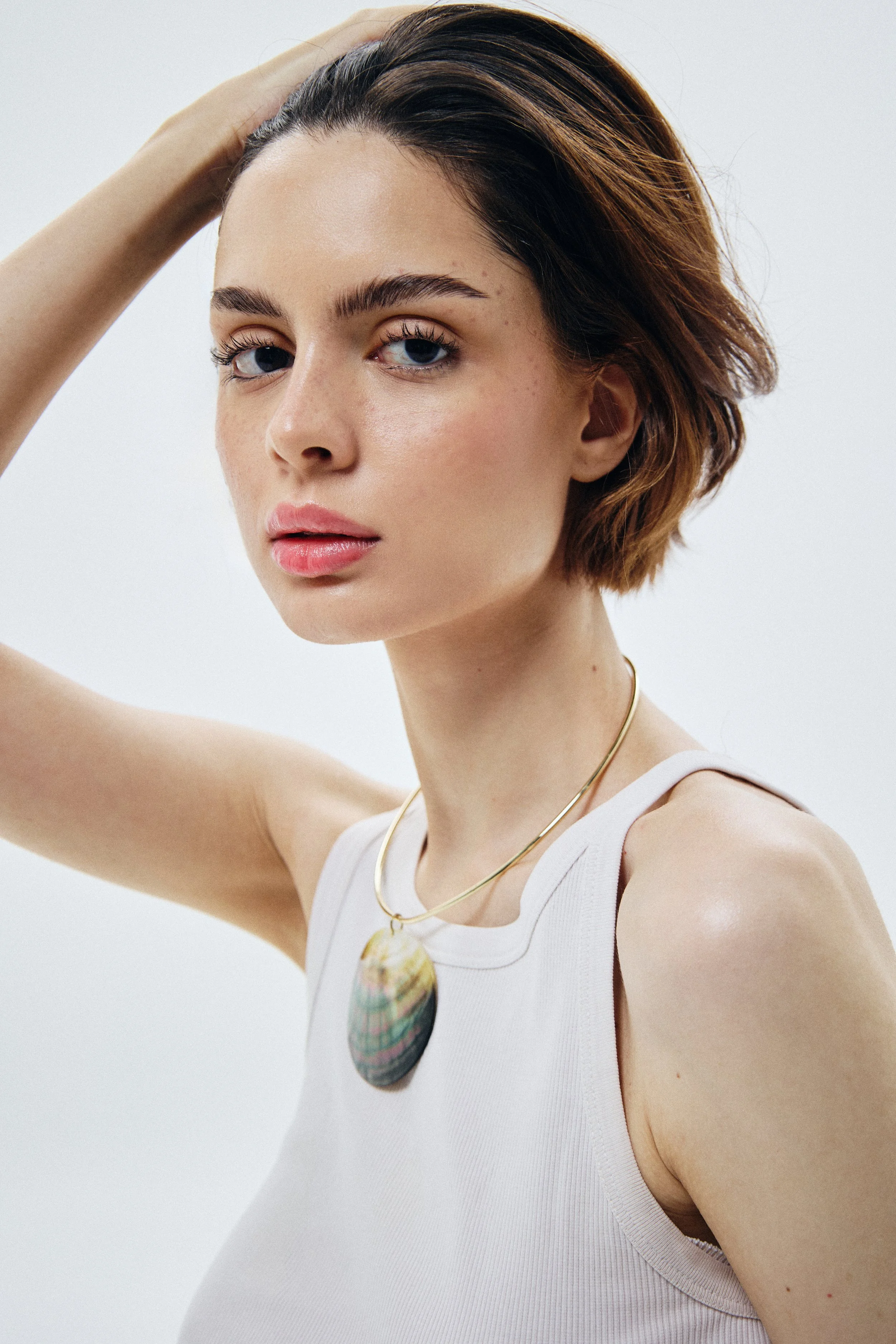 Close-up of a young woman with short brown hair, blue eyes, and freckles, wearing a white tank top, gold necklaces, and touching her hair with her left hand, against a plain white background.
