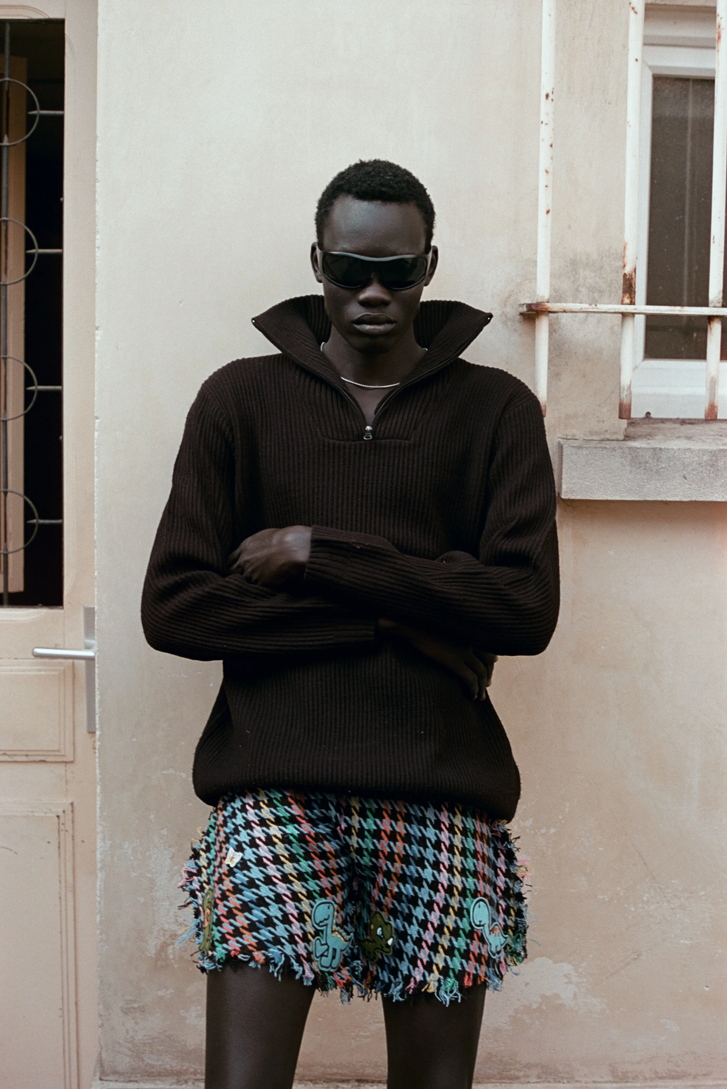A person with dark skin and short curly hair wearing a dark brown sweater with a high collar, sunglasses, a silver necklace, and colorful patterned shorts with cartoon characters, standing against a beige wall with a window and iron bars.