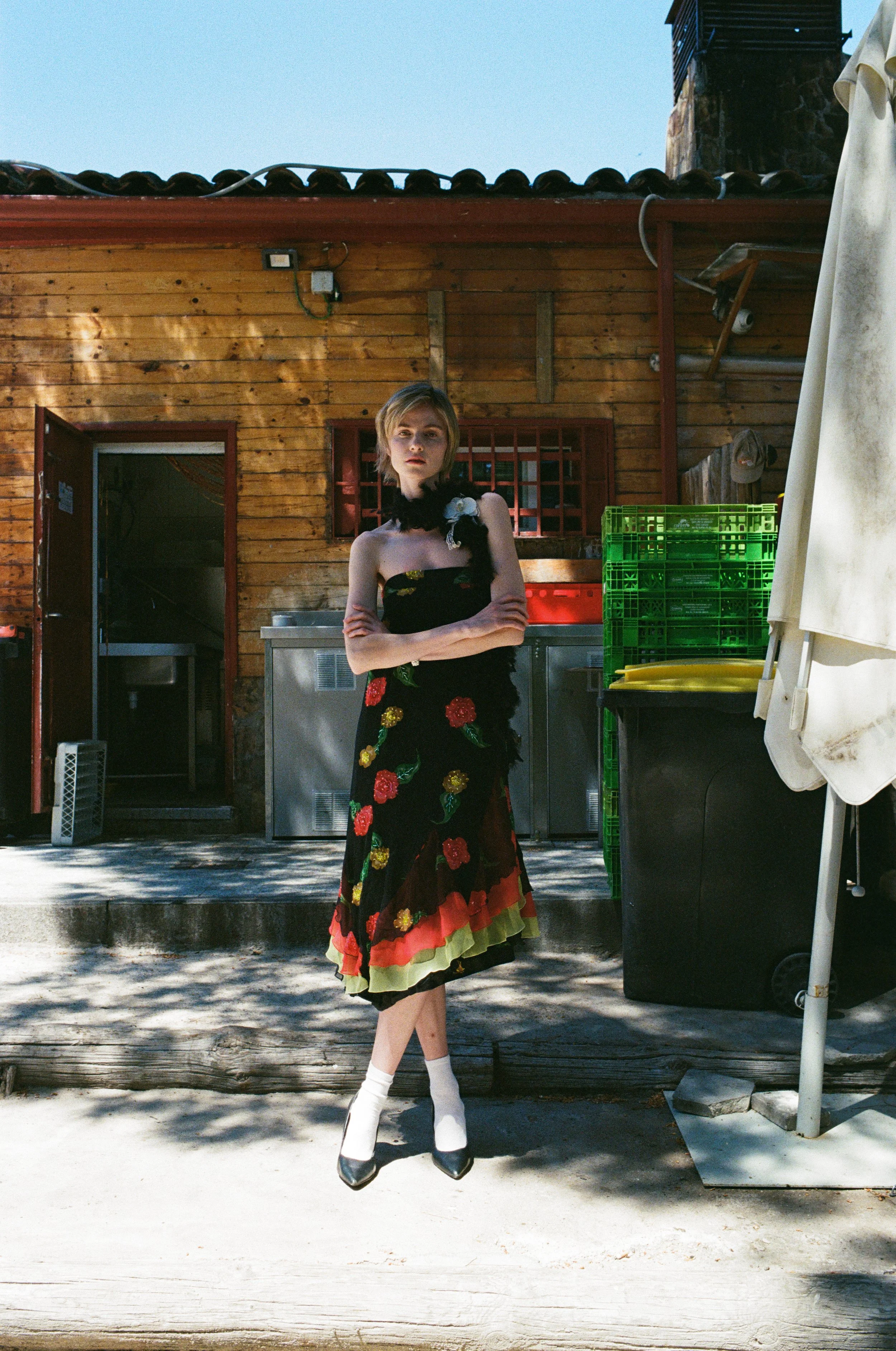 A woman stands outdoors in front of a wooden building with her arms crossed. She wears a strapless dress with a floral pattern and tiered skirt, knee-high white socks, and black heels. Shadows of trees create patterns on the sidewalk.