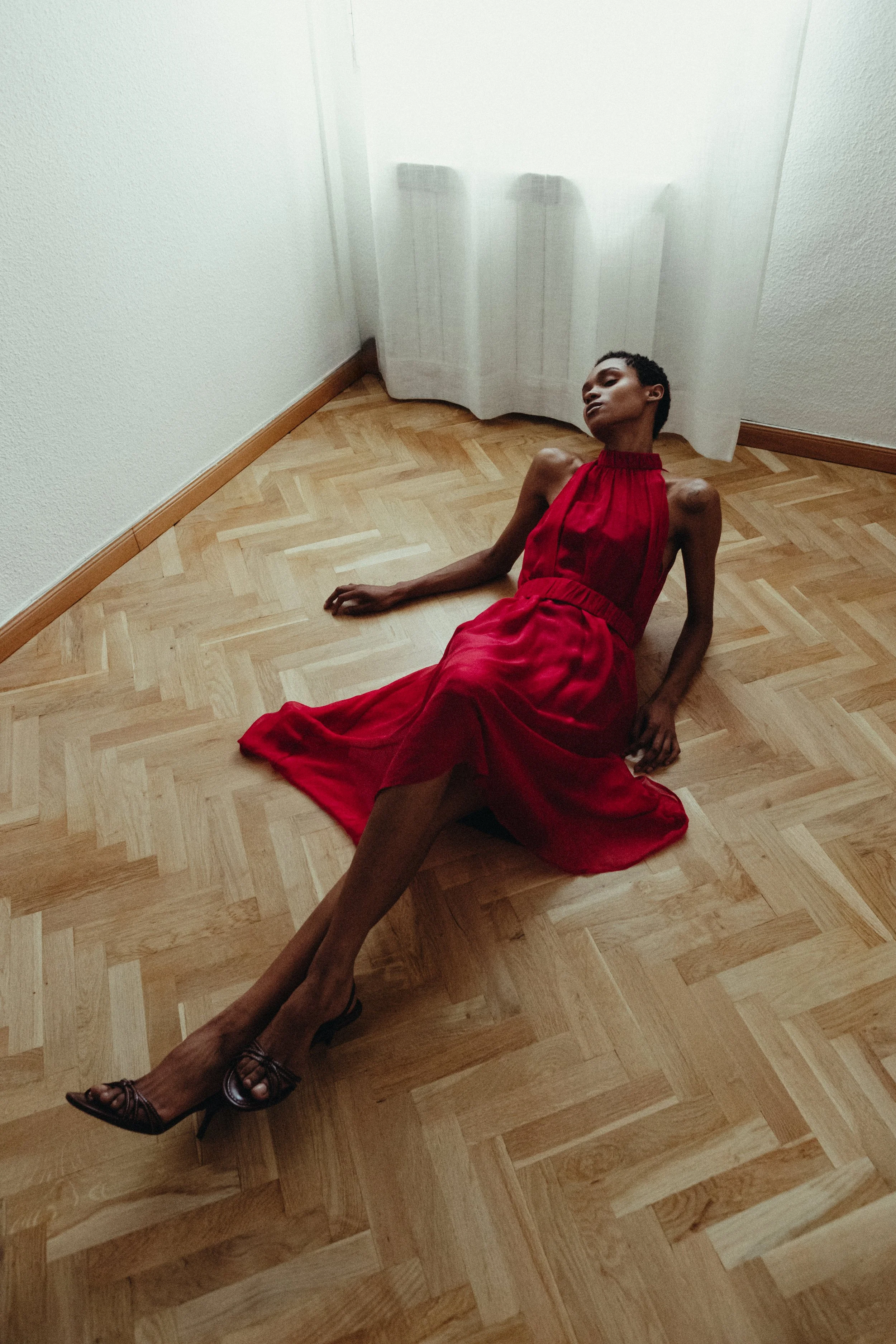 A person with dark skin and short curly hair is sitting on a wooden floor next to a white wall with a curtain. They are wearing a red sleeveless dress and black high-heeled shoes, with one leg extended and the other bent, eyes closed, and head tilted