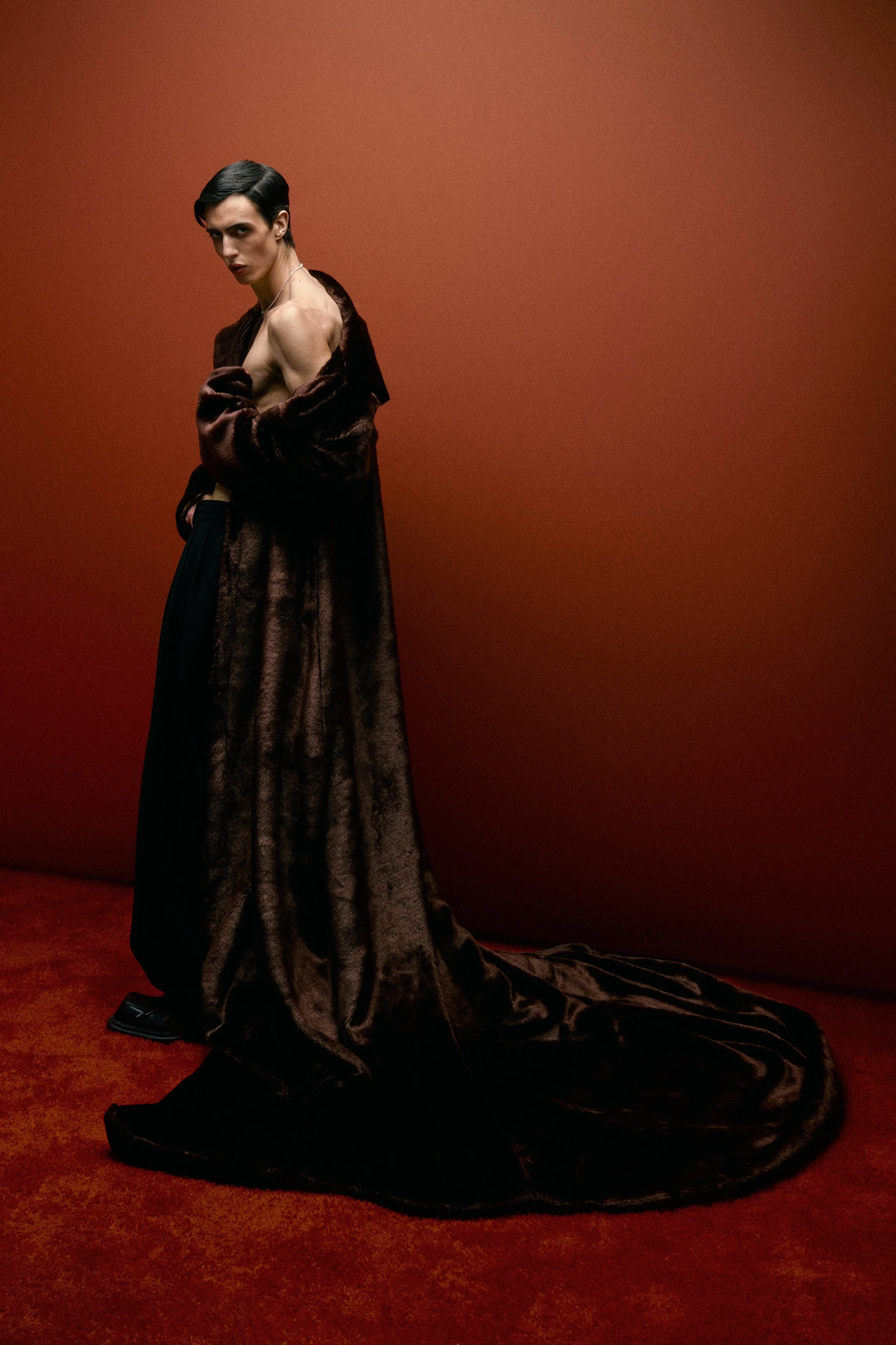 A person with short dark hair and makeup, wearing a long, dark brown fur coat, draped over their shoulders, with no shirt underneath, standing against a reddish-brown background with a shadow cast on the wall.