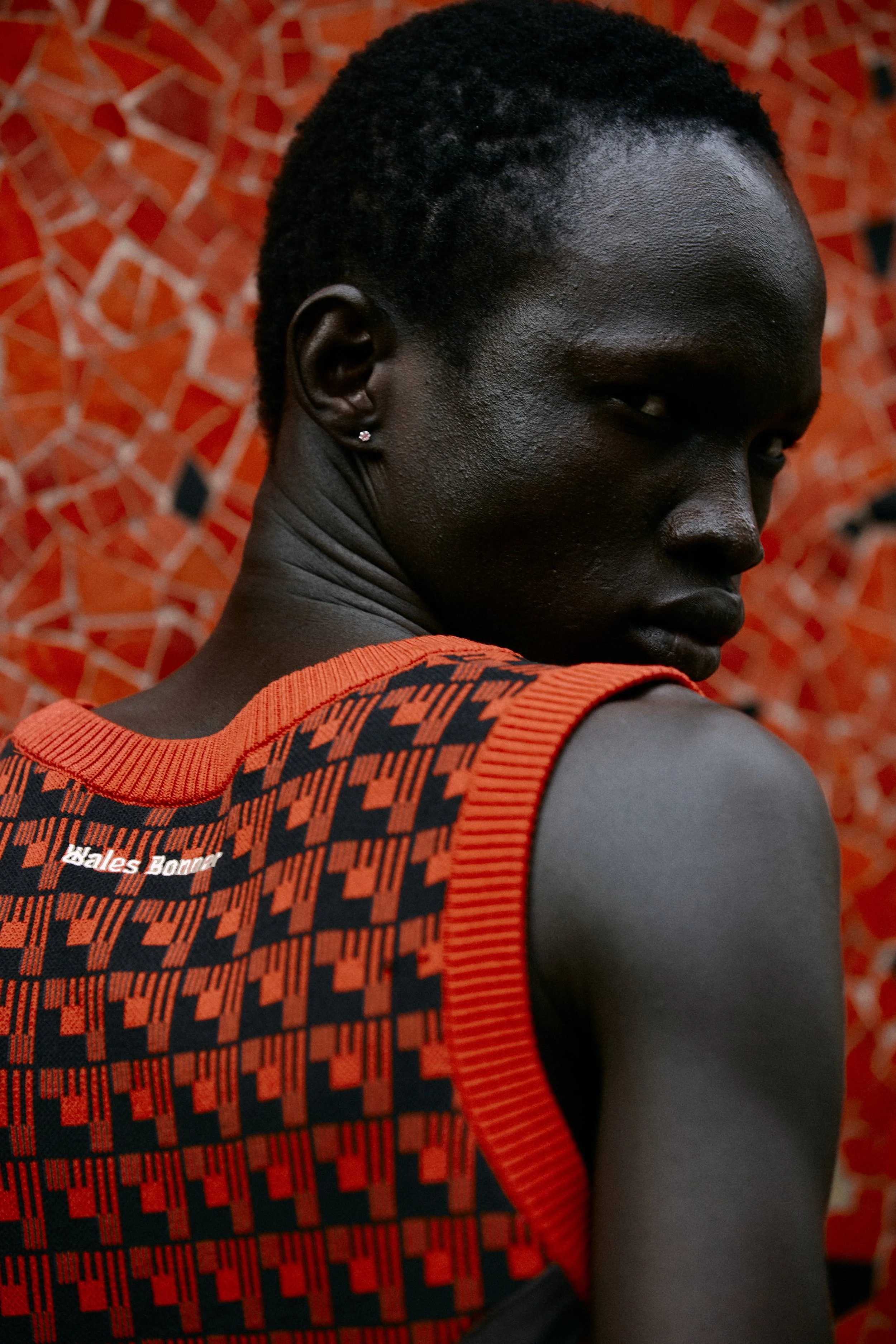 A close-up of a person with short curly black hair, dark skin, wearing a red and black sports jersey with the text 'Wales Bomber' and an orange background with a geometric pattern.