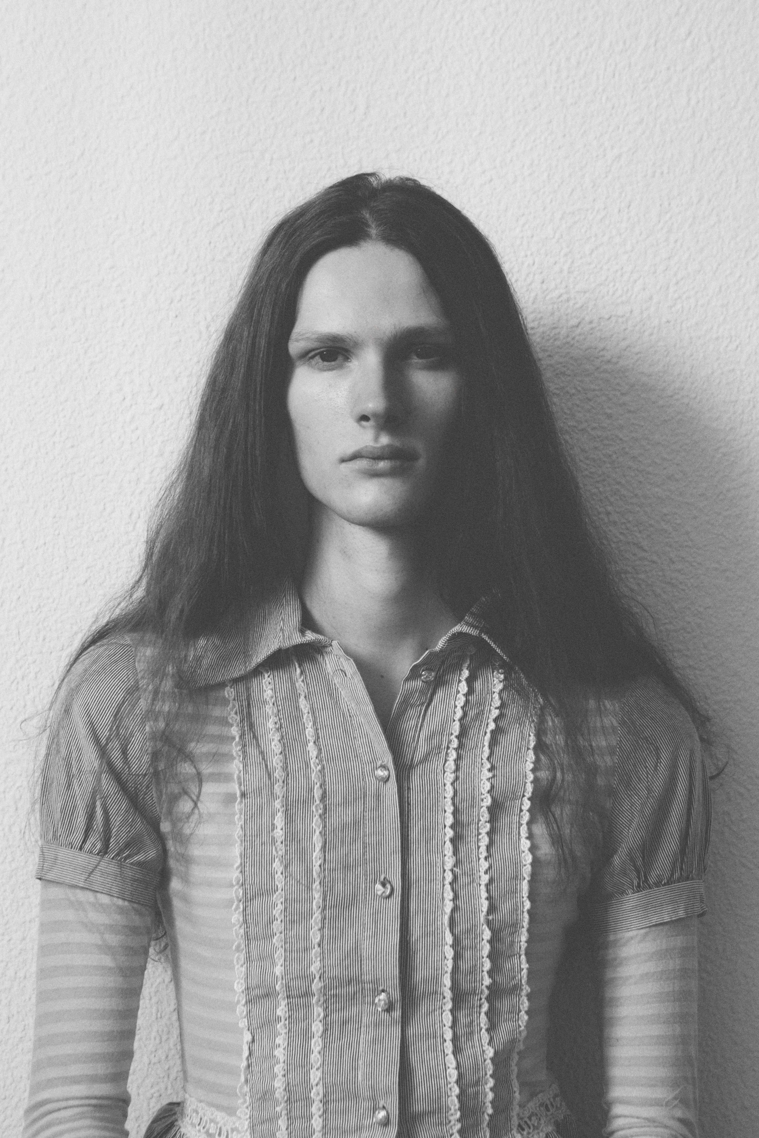 Black and white photo of a woman with long dark hair, wearing a striped shirt with lace details, standing against a plain wall.