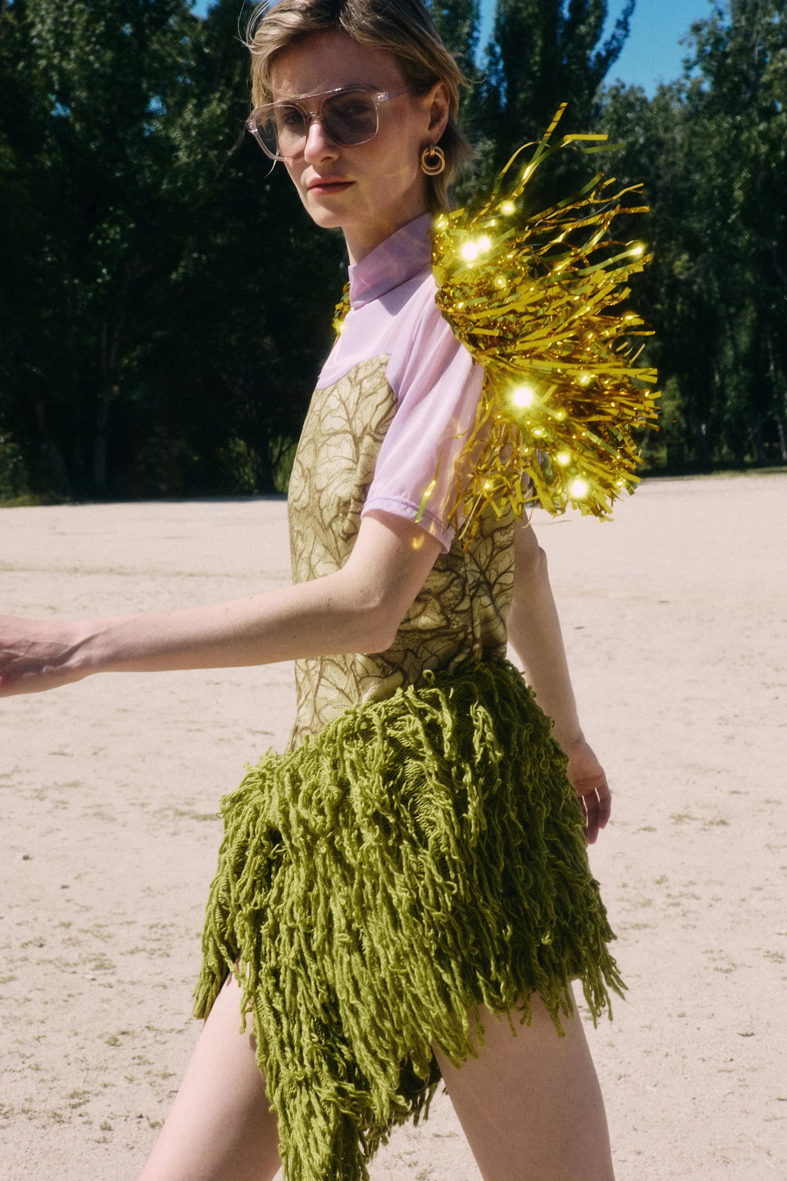 A woman wearing sunglasses and earrings stands outdoors on a sunny day. She is dressed in a pink top with a gold patterned overlay and a green, textured skirt. She has a large gold and black shoulder accessory that appears to be adorned with lights o