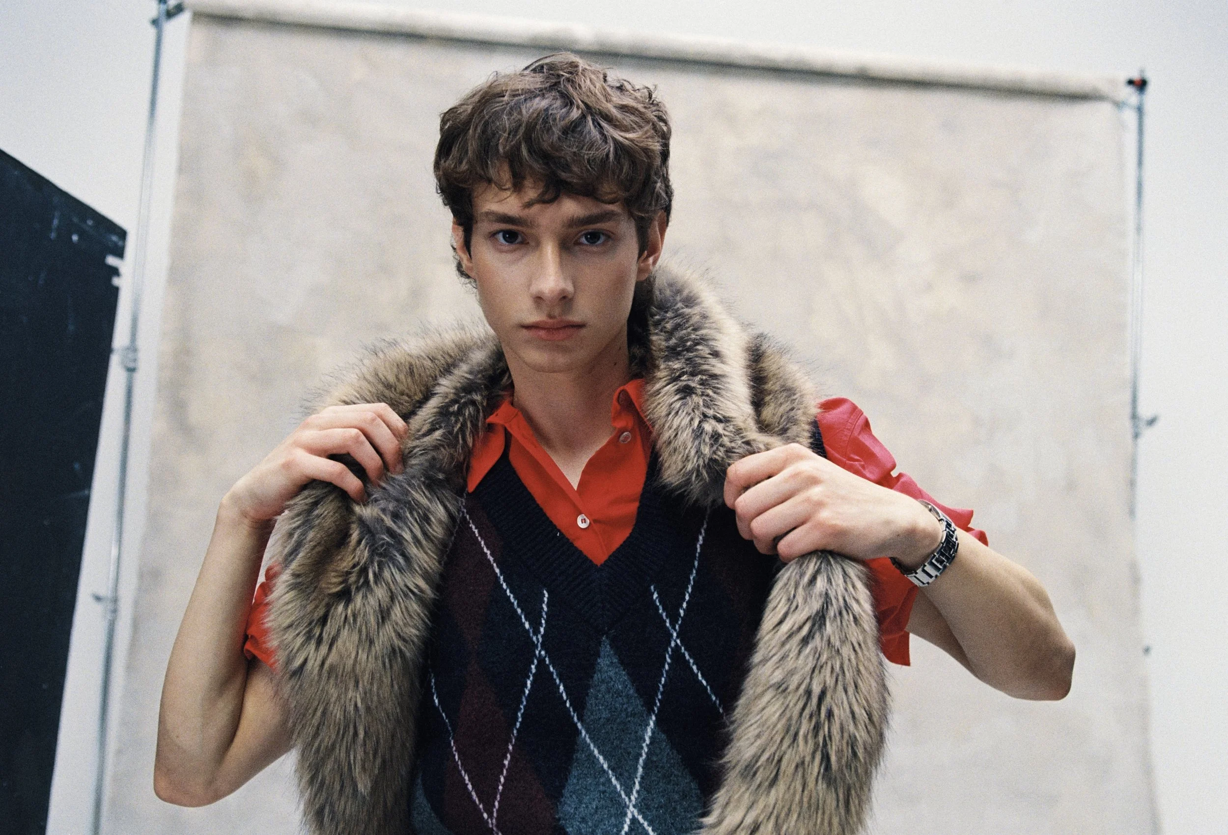 A young man with curly hair wearing a red shirt, navy sweater vest with an argyle pattern, and a fur-lined coat, standing indoors.